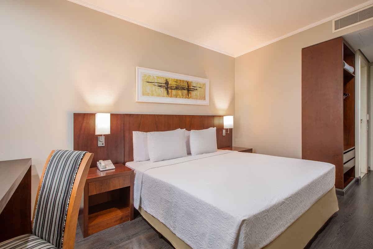 Ein Hotelzimmer mit einem großen Bett, einem hölzernen Kopfteil, zwei Nachttischlampen, einem Nachttisch, einem gestreiften Sessel und einem Bild an der Wand.
