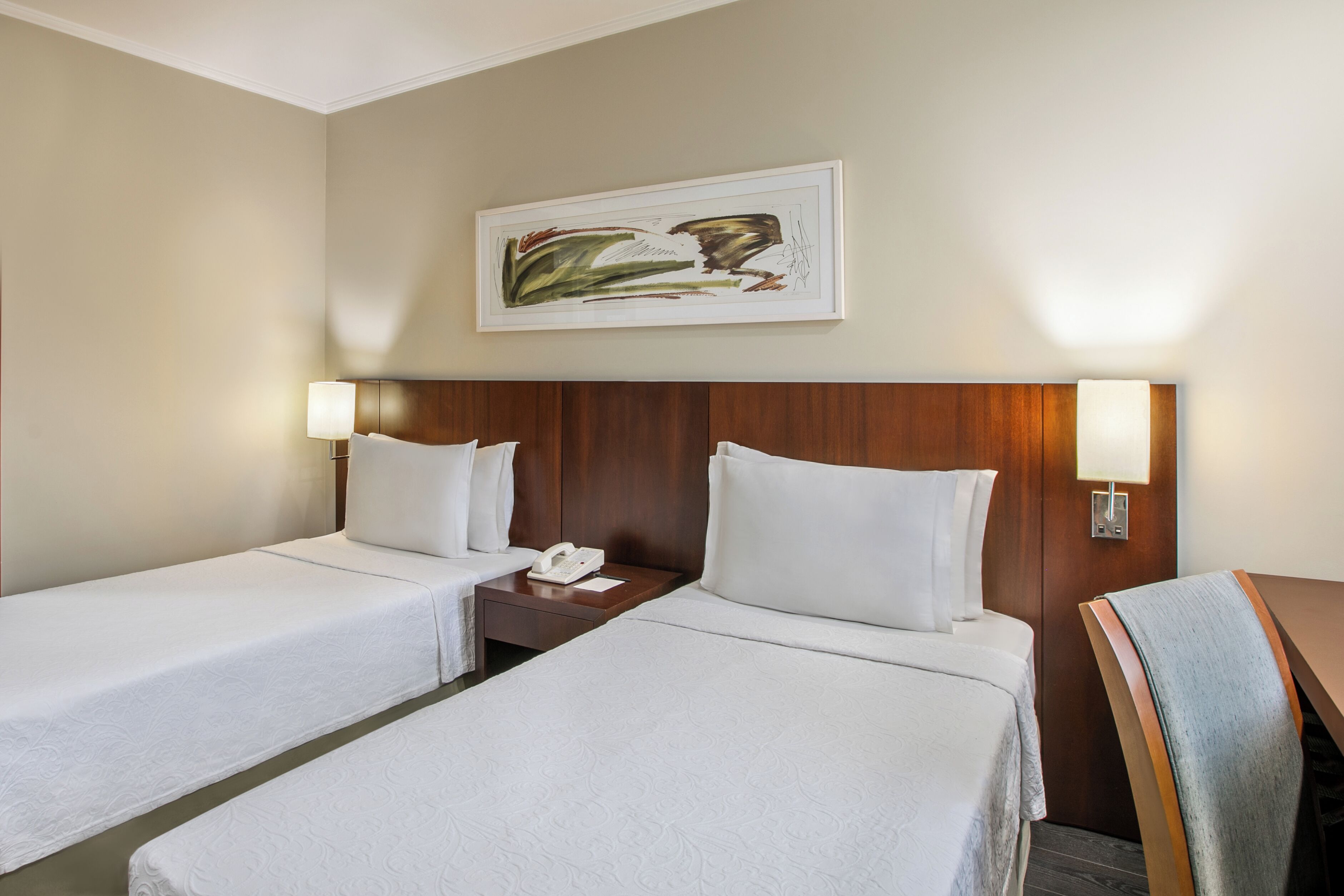 A hotel room with two single beds, a wooden headboard, a painting above the beds, and bedside lamps.