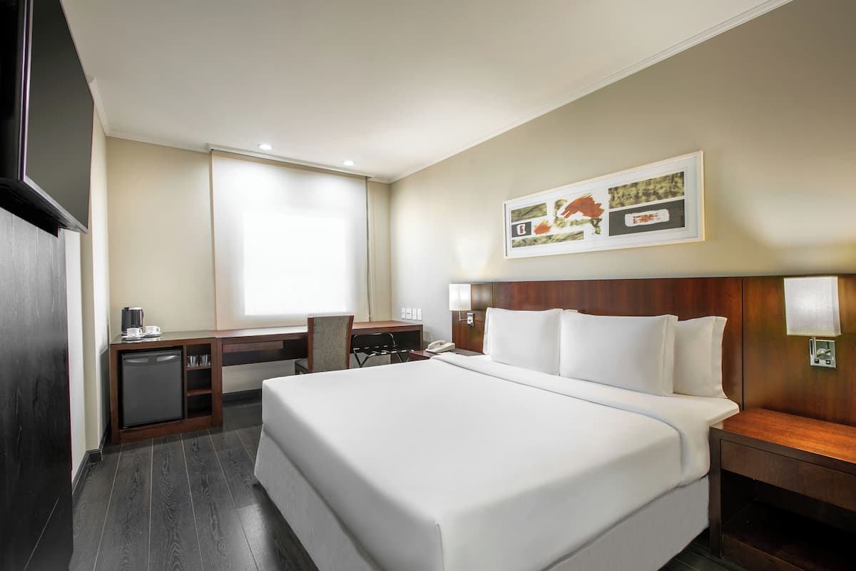 Ein Hotelzimmer mit einem großen Bett, einem Schreibtisch mit Computer, einer kleinen Kühlbox und einem Fenster mit Vorhängen.