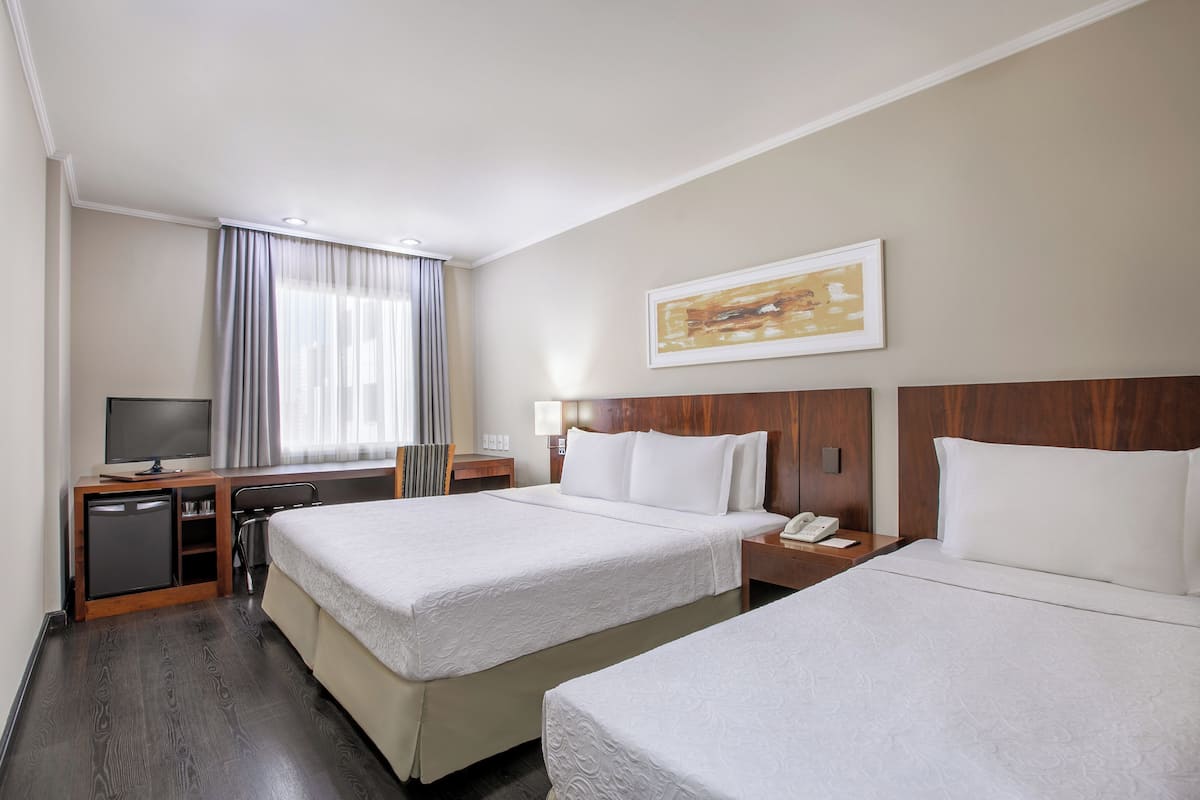 Ein Hotelzimmer mit zwei Betten, einem Schreibtisch mit Fernseher, einem kleinen Kühlschrank und einem Fenster mit Vorhängen.