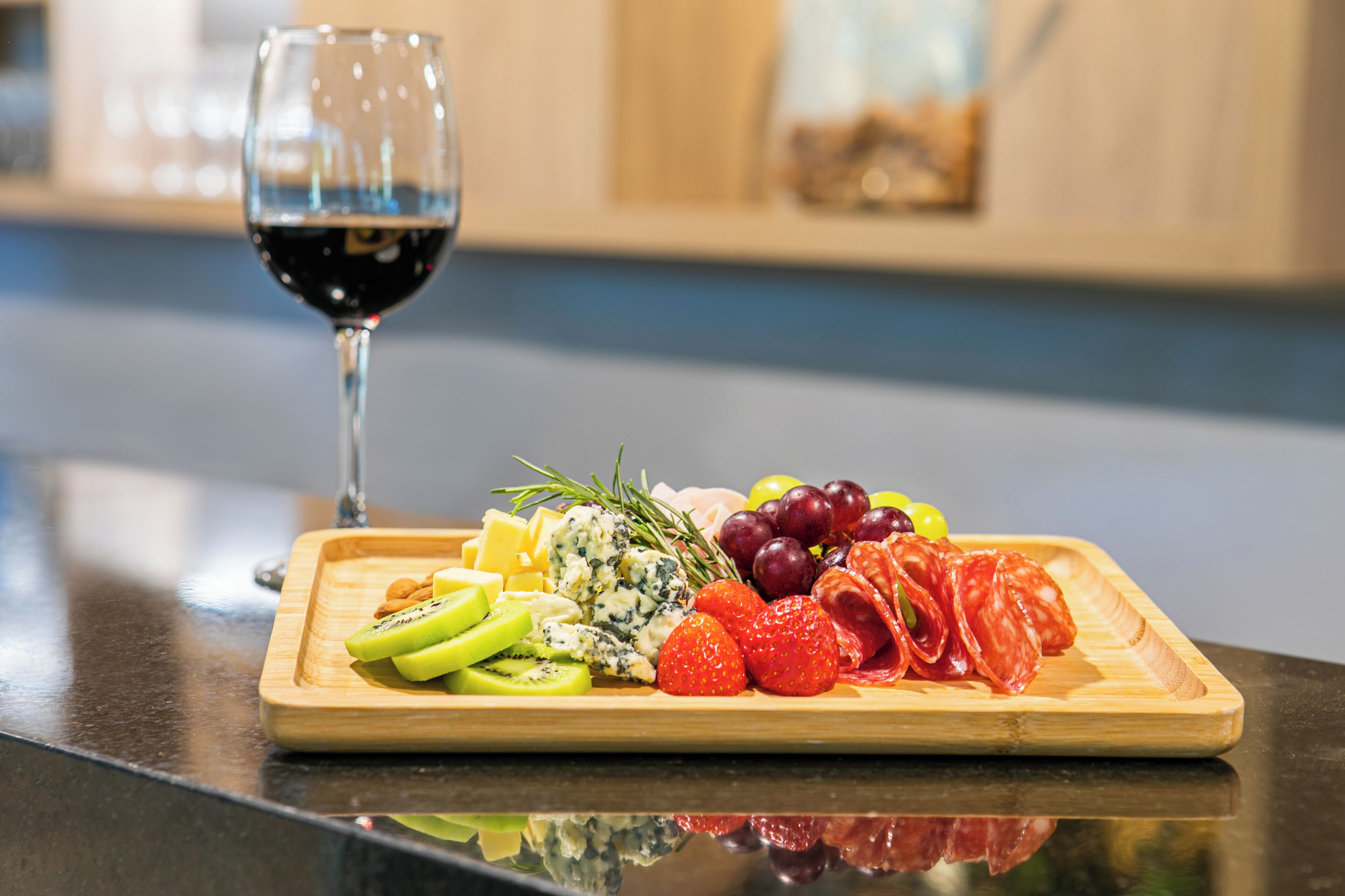A wooden board with various fruits, cheese, and meats, accompanied by a glass of red wine on a reflective surface.