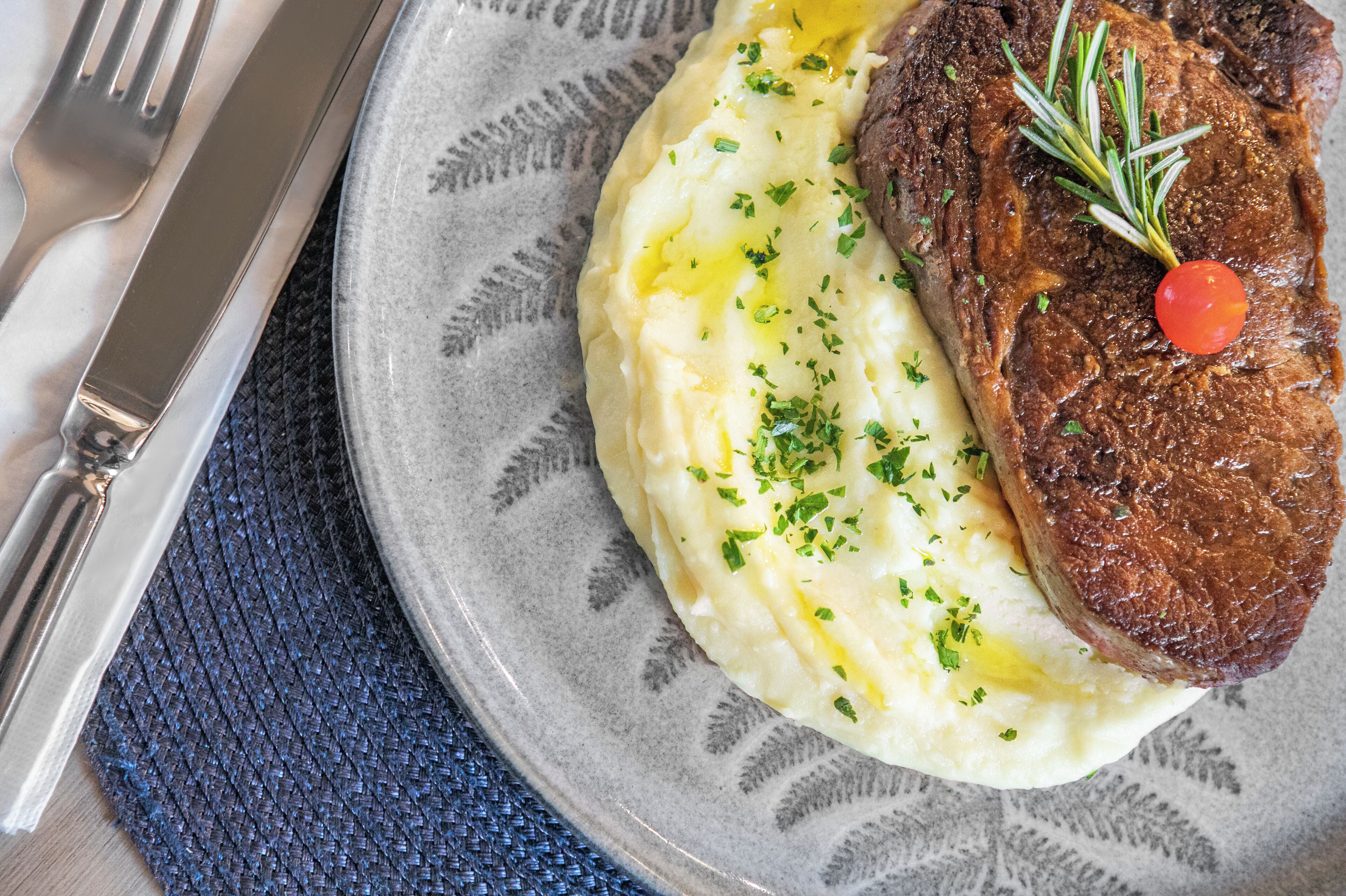 Un plato con puré de papas, un bistec adornado con hierbas y un tomate cherry.