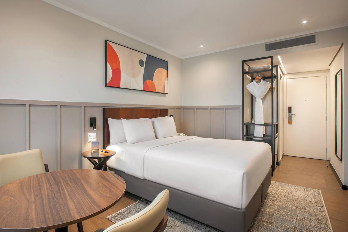 Ein Hotelzimmer mit einem Bett, einem runden Holztisch, einem Sessel, einem Nachttisch, einem Wandbild und einem Spiegel.