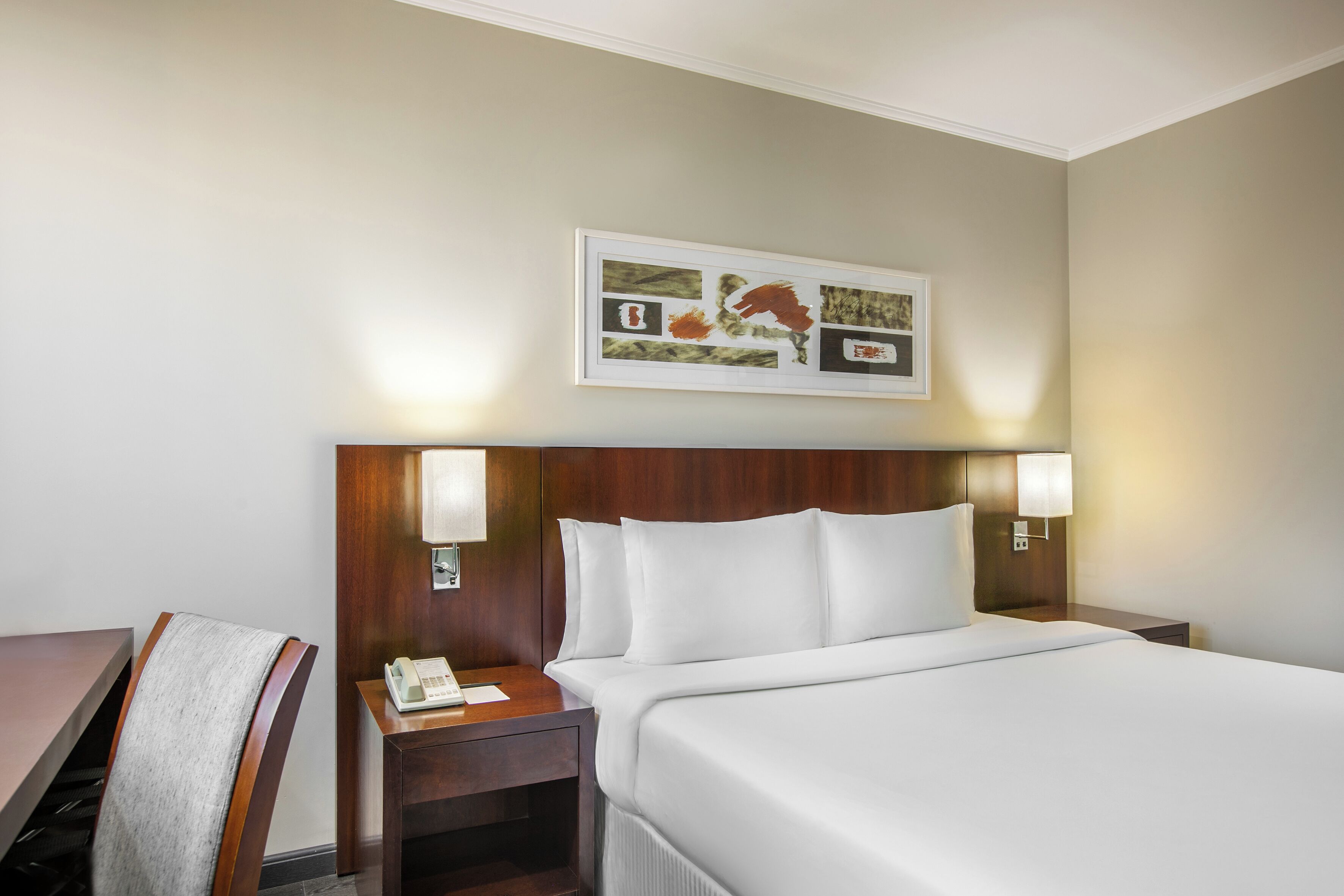 Una habitación de hotel con cama, lámparas de noche, escritorio, silla y un cuadro en la pared.