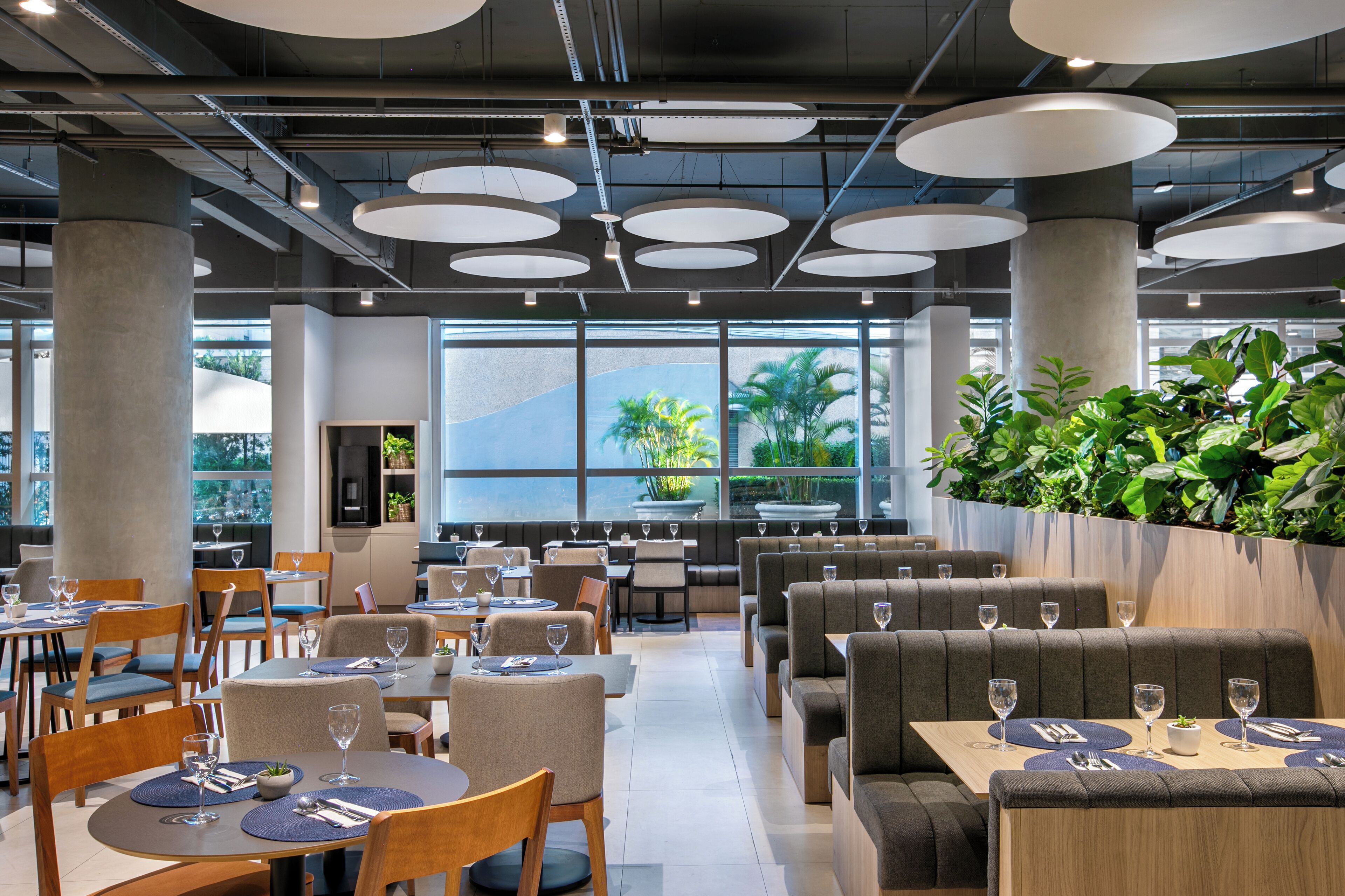 Ein modernes Restaurant mit runden Tischen, Sitzbänken und einem großen Fenster mit Blick auf Grünflächen.
