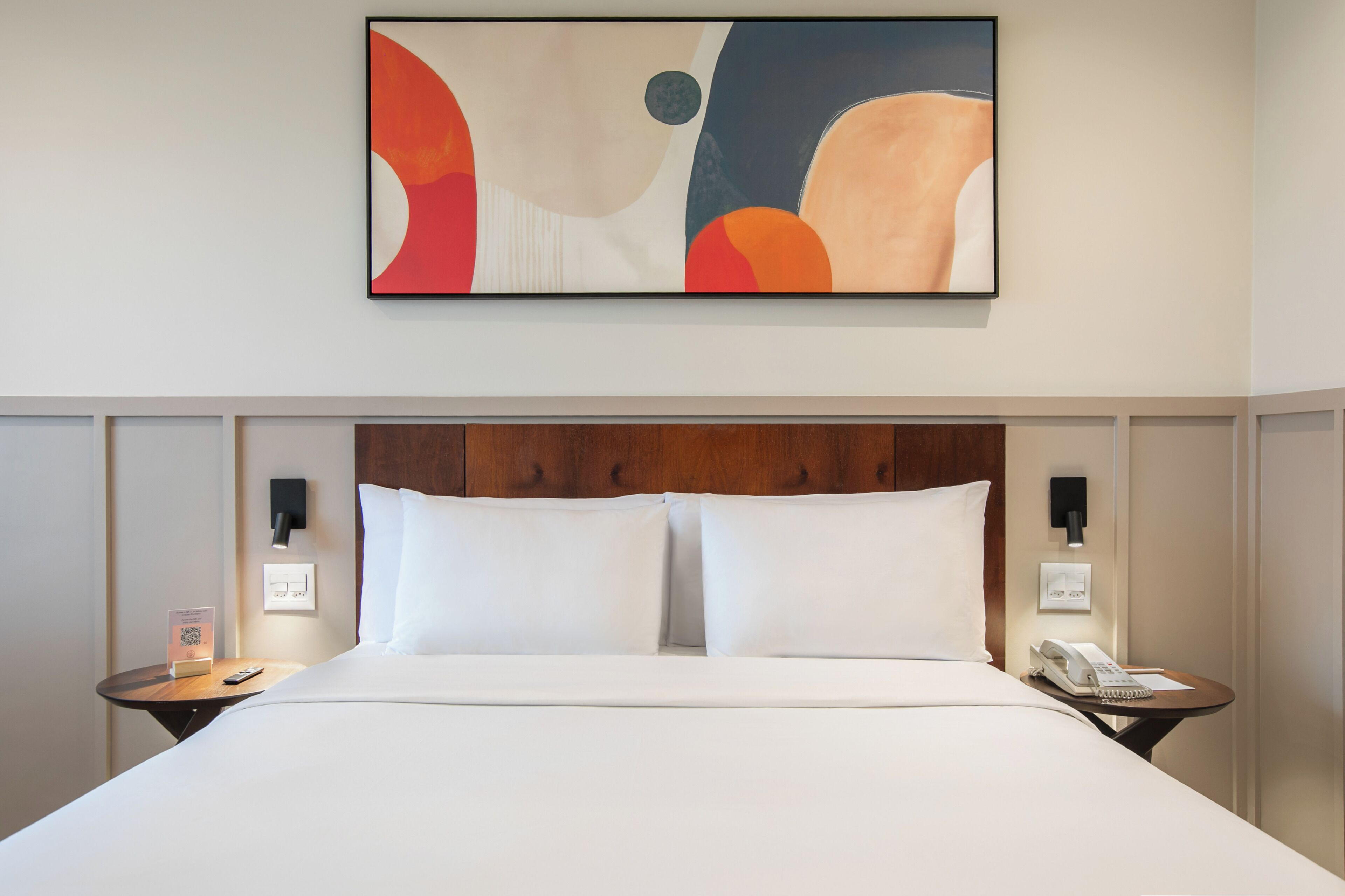 Quarto de hotel com uma cama grande, duas mesas de cabeceira, uma obra de arte na parede e uma cabeceira de madeira.