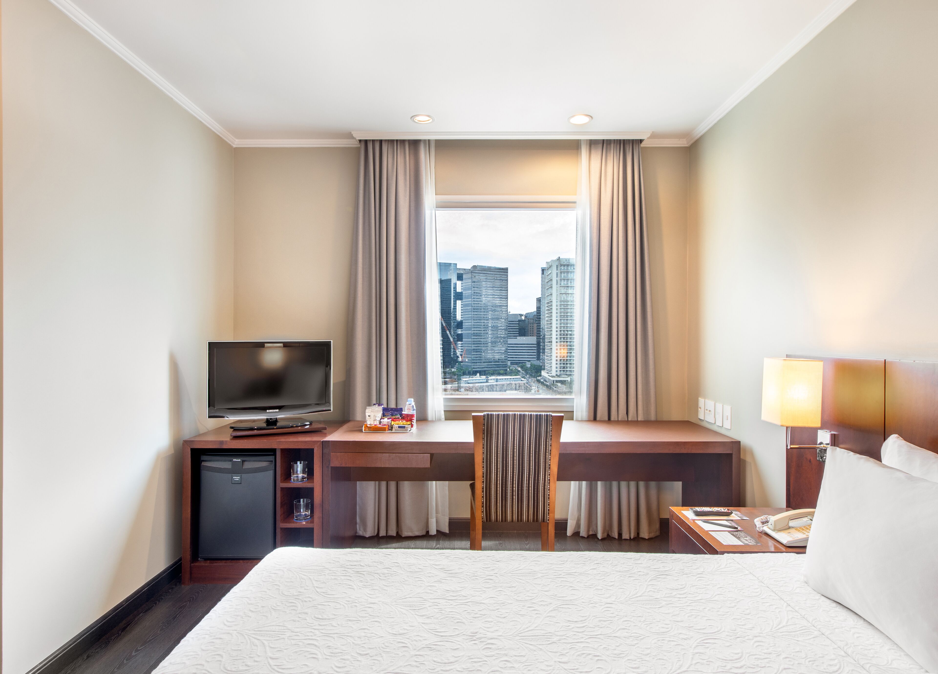 A hotel room with a large bed, a desk with a TV, a small fridge, a telephone, and a window with curtains.