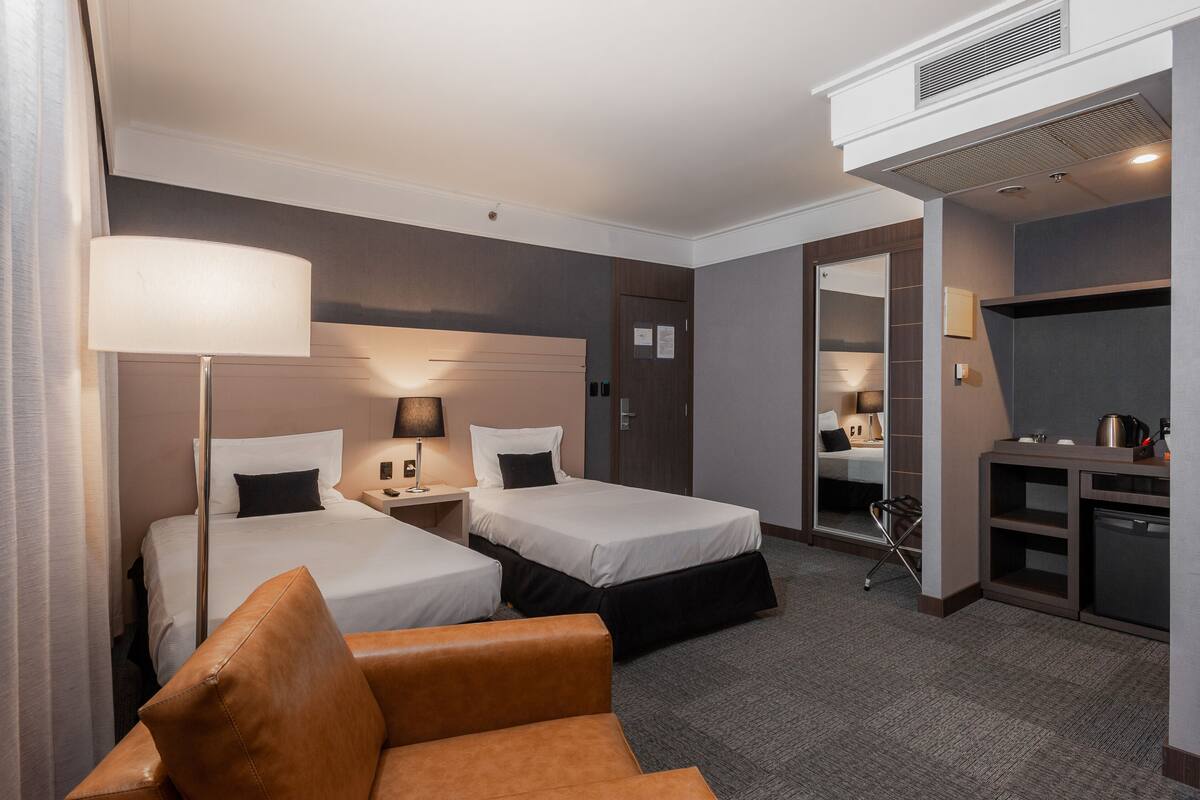 A hotel room with two beds, a desk, a chair, a kitchenette, and a bathroom visible through an open door.