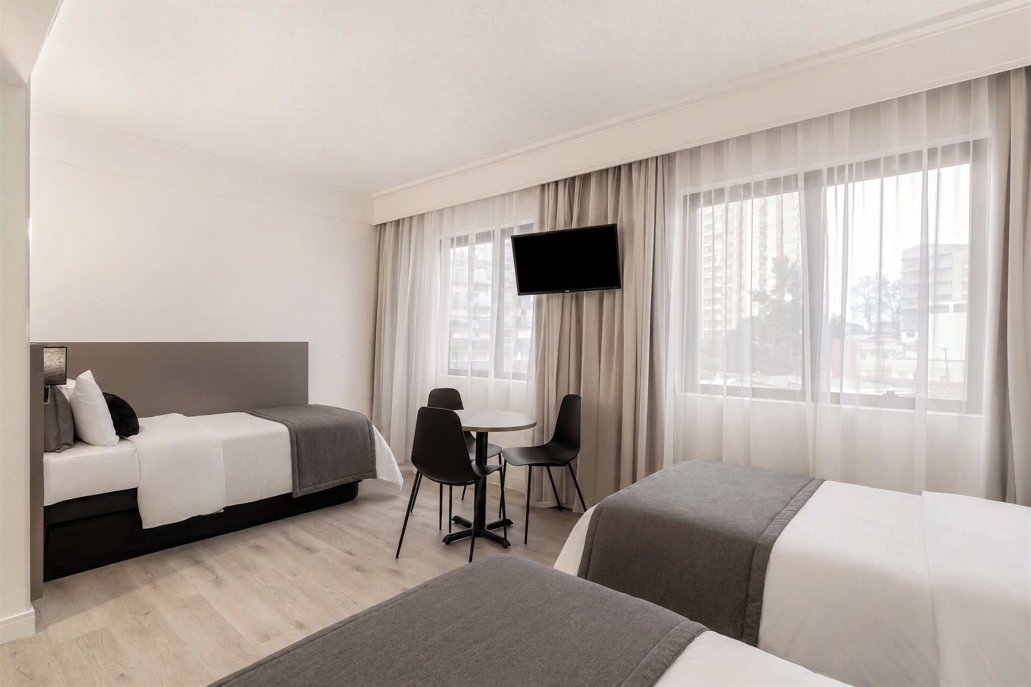 Habitació d'hotel amb dos llits, una taula de treball, cadires, televisió i finestres grans amb cortines.