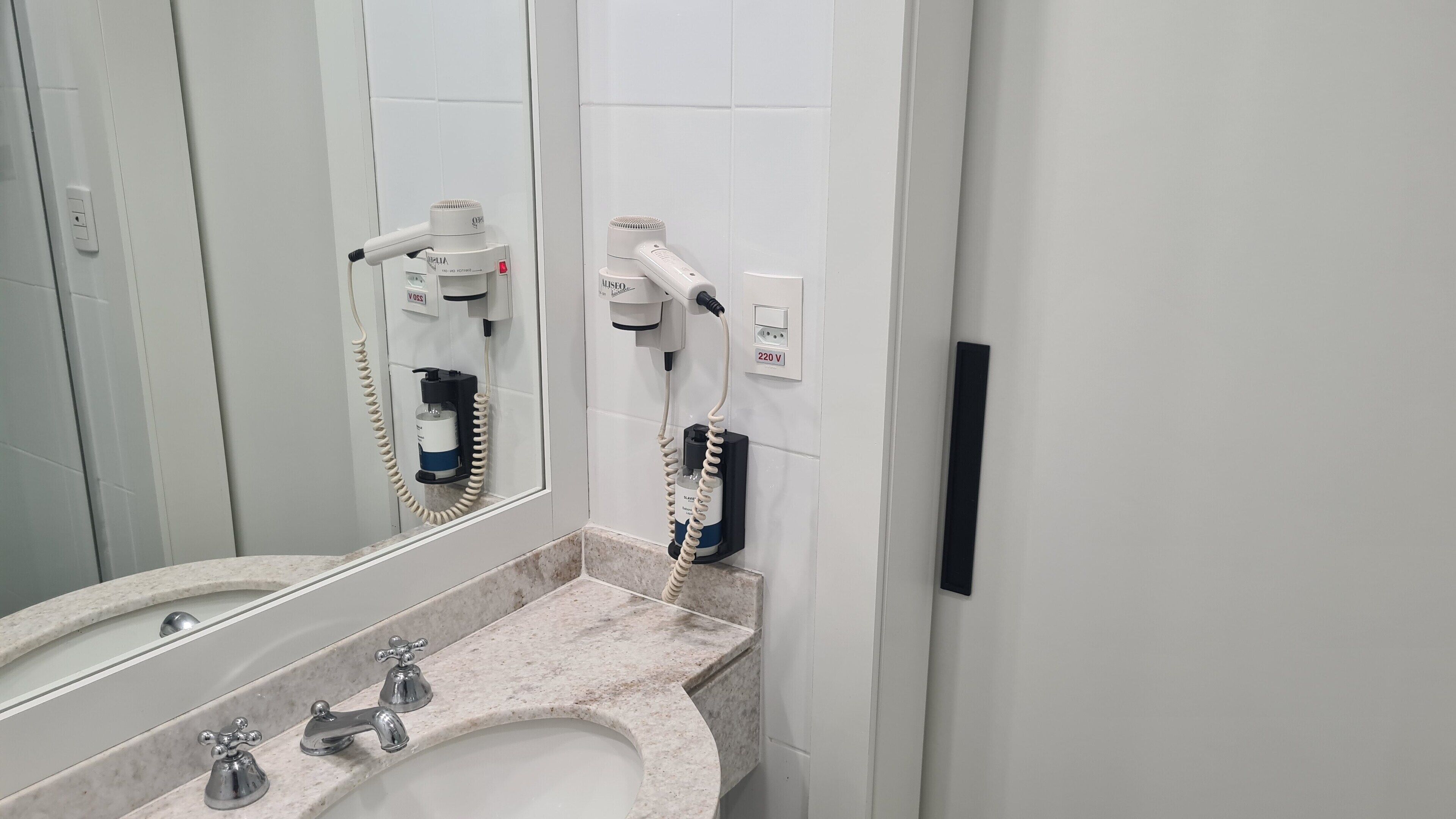 A bathroom with a sink, two mirrors, a hairdryer, and a wall-mounted hairdryer.