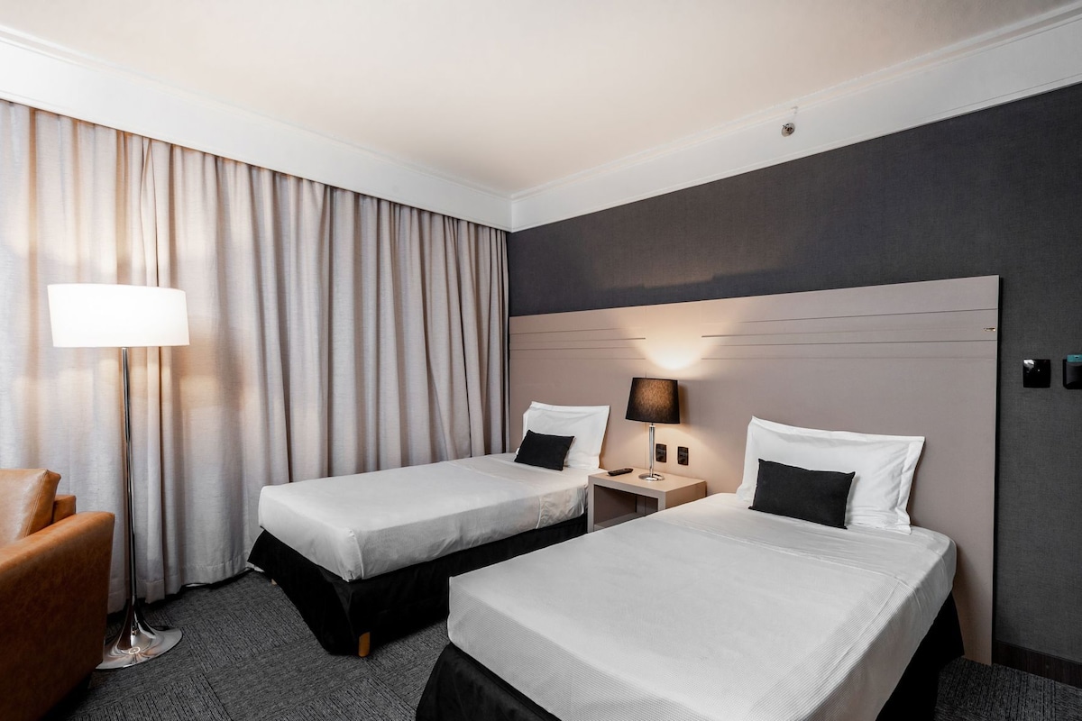 A hotel room with two beds, a desk, a chair, and a lamp.