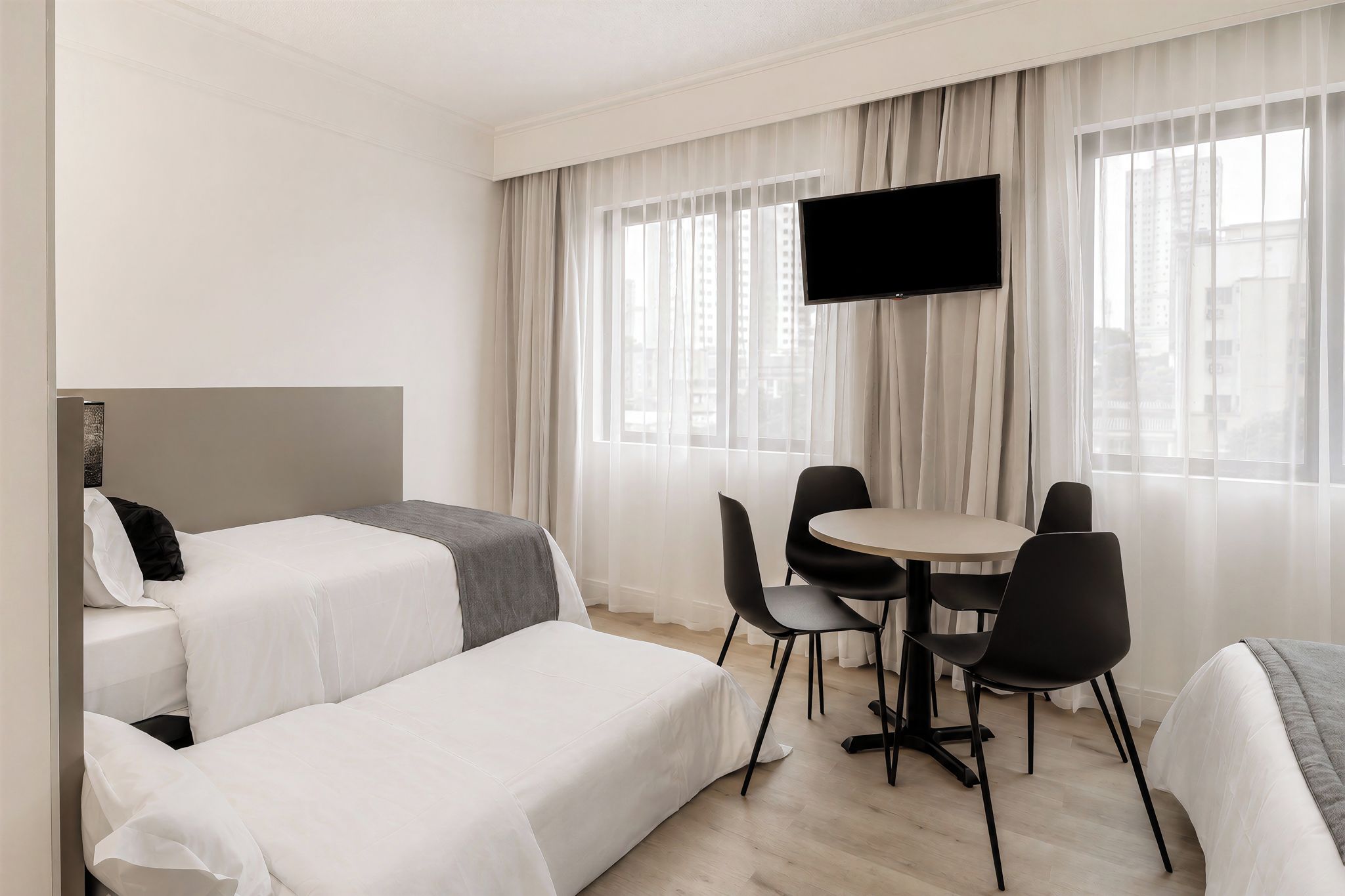 Camera d'albergo con due letti, televisione, un tavolo da pranzo con sedie e vista sui palazzi attraverso la finestra.