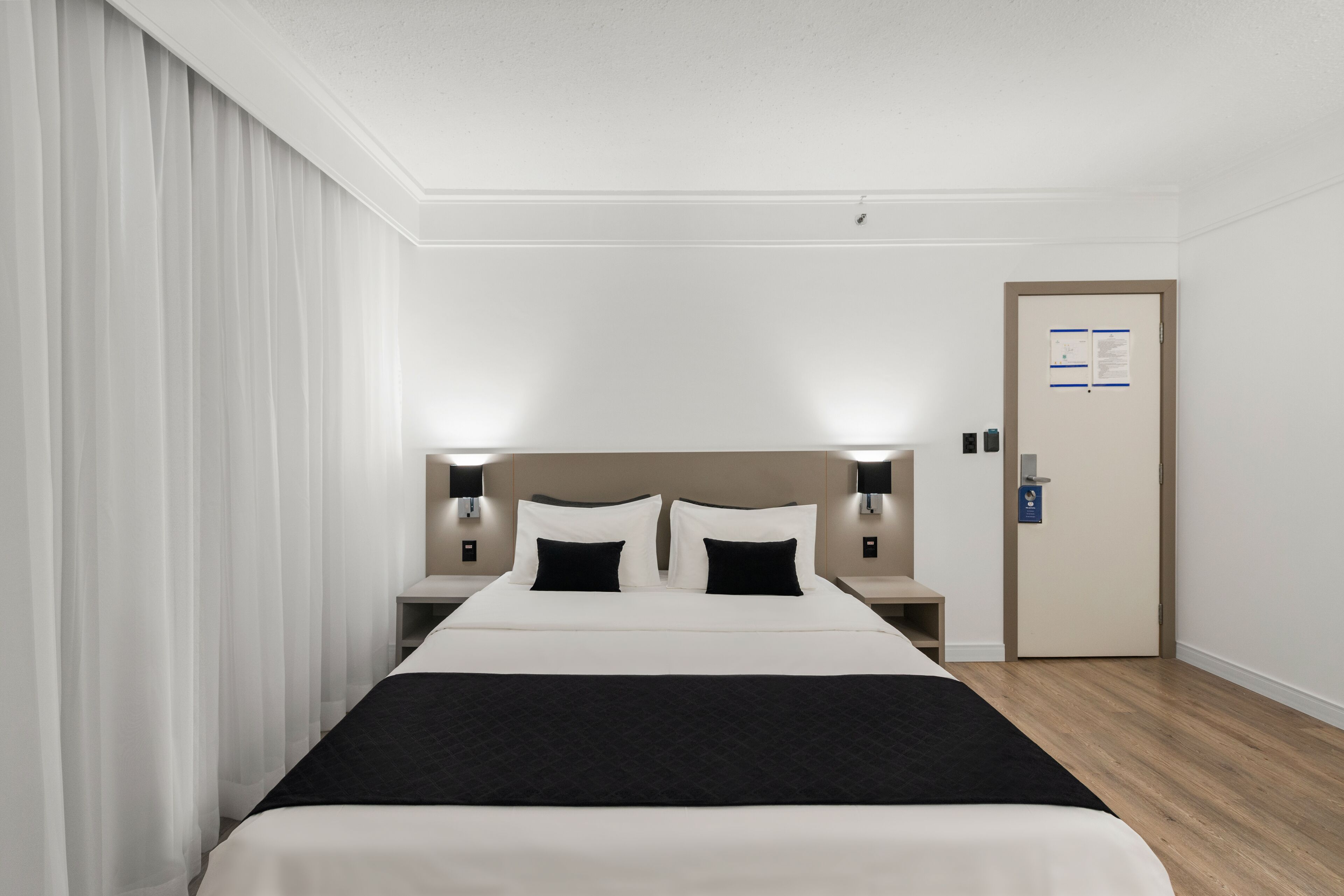 A hotel room with a large bed, bedside tables with lamps, a door with a notice, and a curtain on the left.