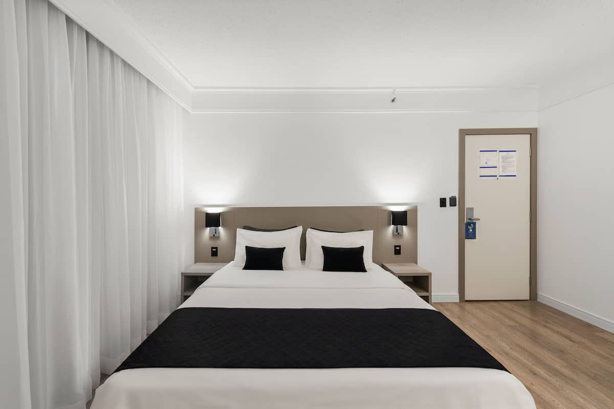 A hotel room with a large bed, bedside tables with lamps, a door with a notice, and a curtain on the left.