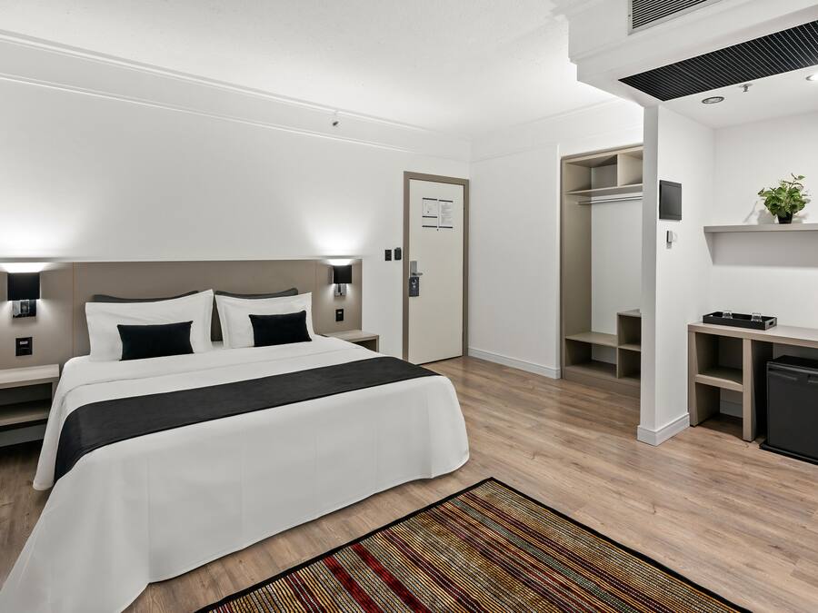 A hotel room with a bed, bedside tables, a desk, a mirror, a small kitchen area, and a door.