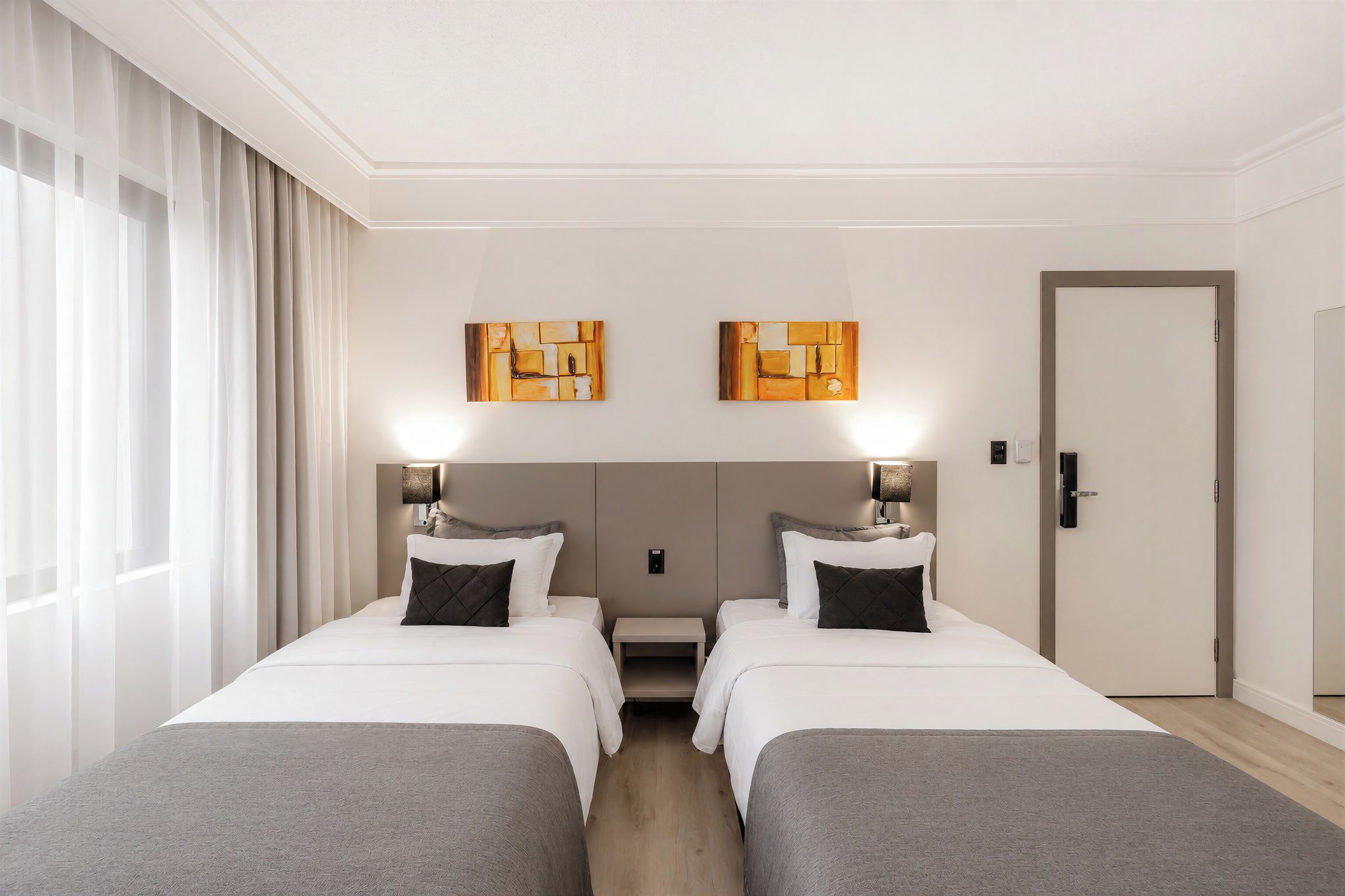 Quarto de hotel com duas camas, uma mesa de cabeceira, uma porta e duas pinturas abstratas na parede.