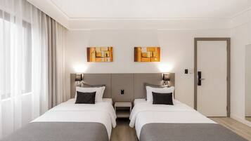 Quarto de hotel com duas camas, uma mesa de cabeceira, uma porta e duas pinturas abstratas na parede.