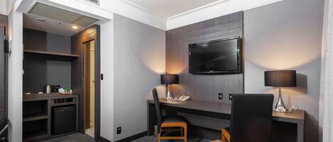 Habitación de hotel con escritorio, silla, televisor y una pequeña kitchenette.