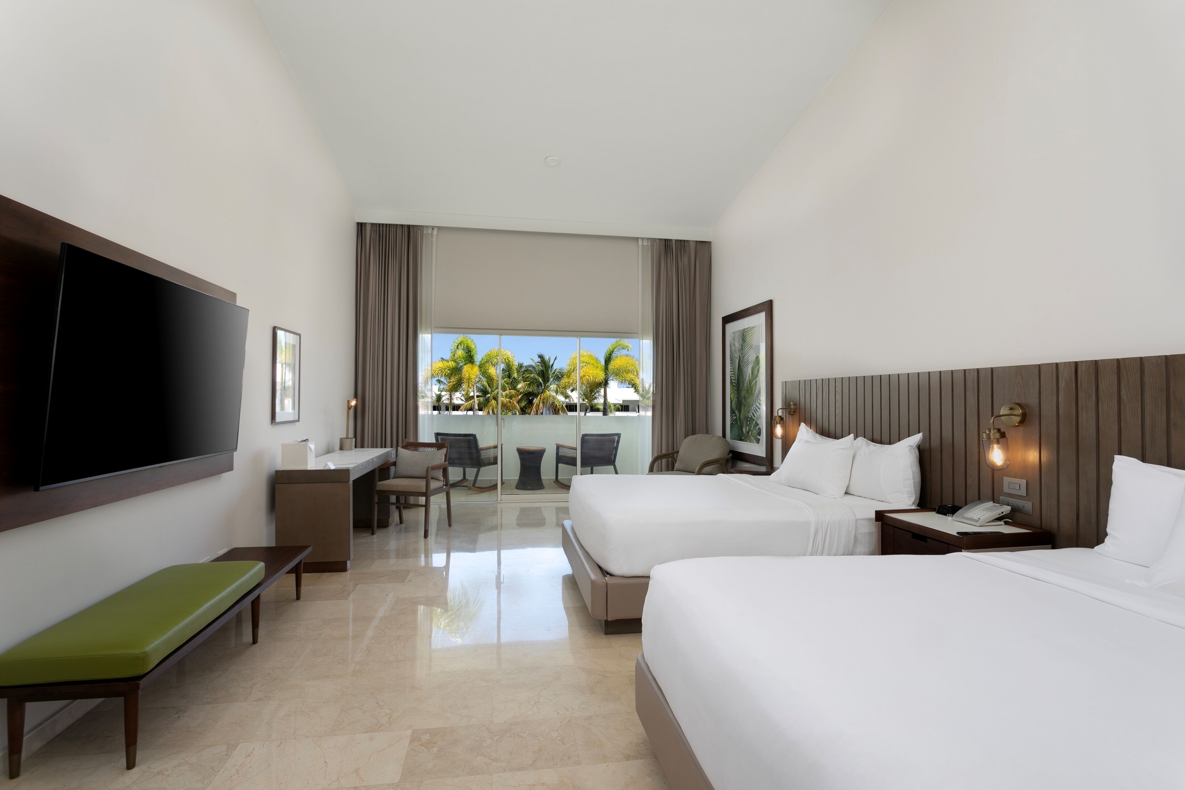 A hotel room with two beds, a TV, a desk, and a balcony with a view of palm trees.