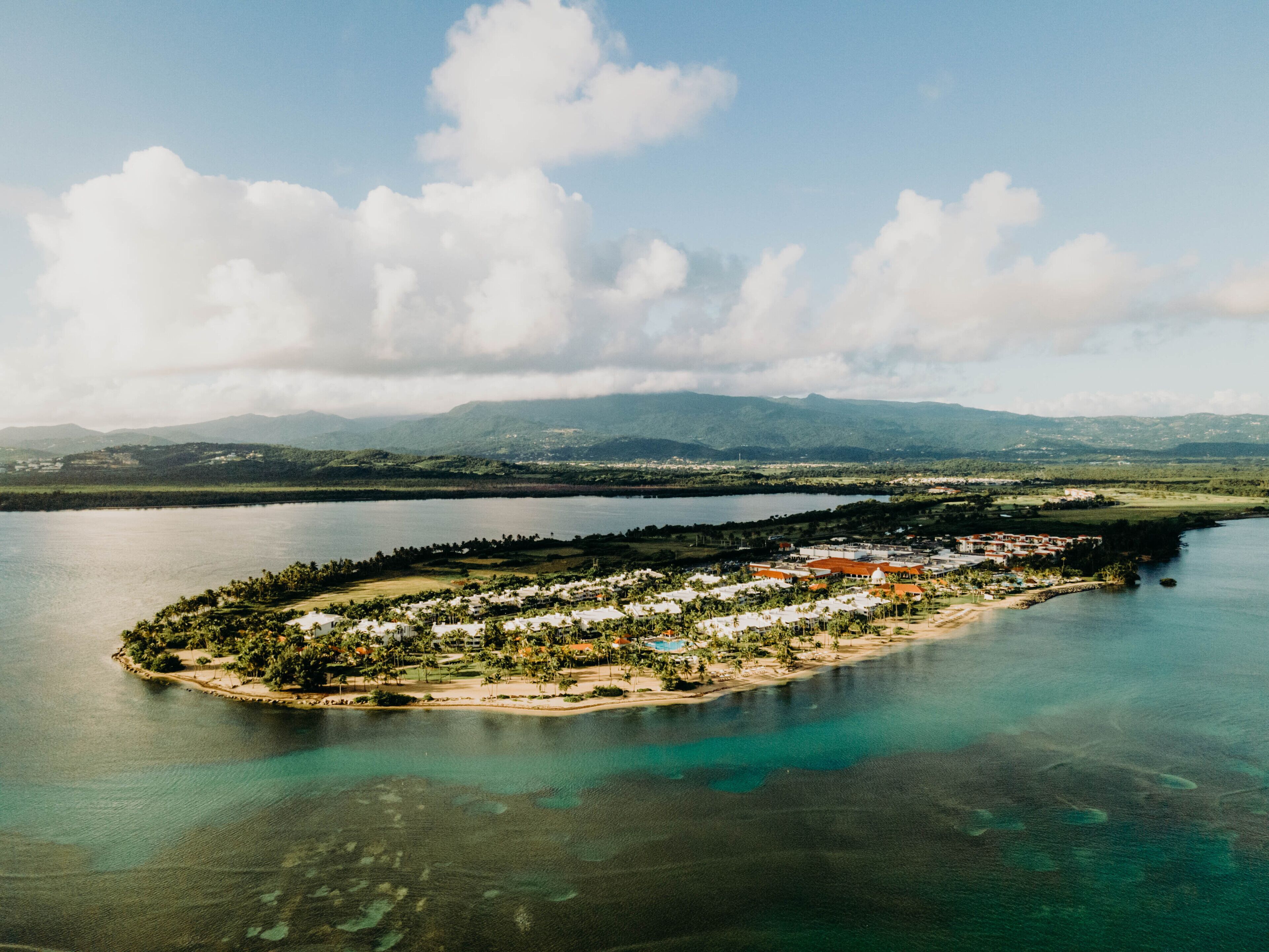 An aerial view of a small island with a resort area, surrounded by water and mountains in the background.