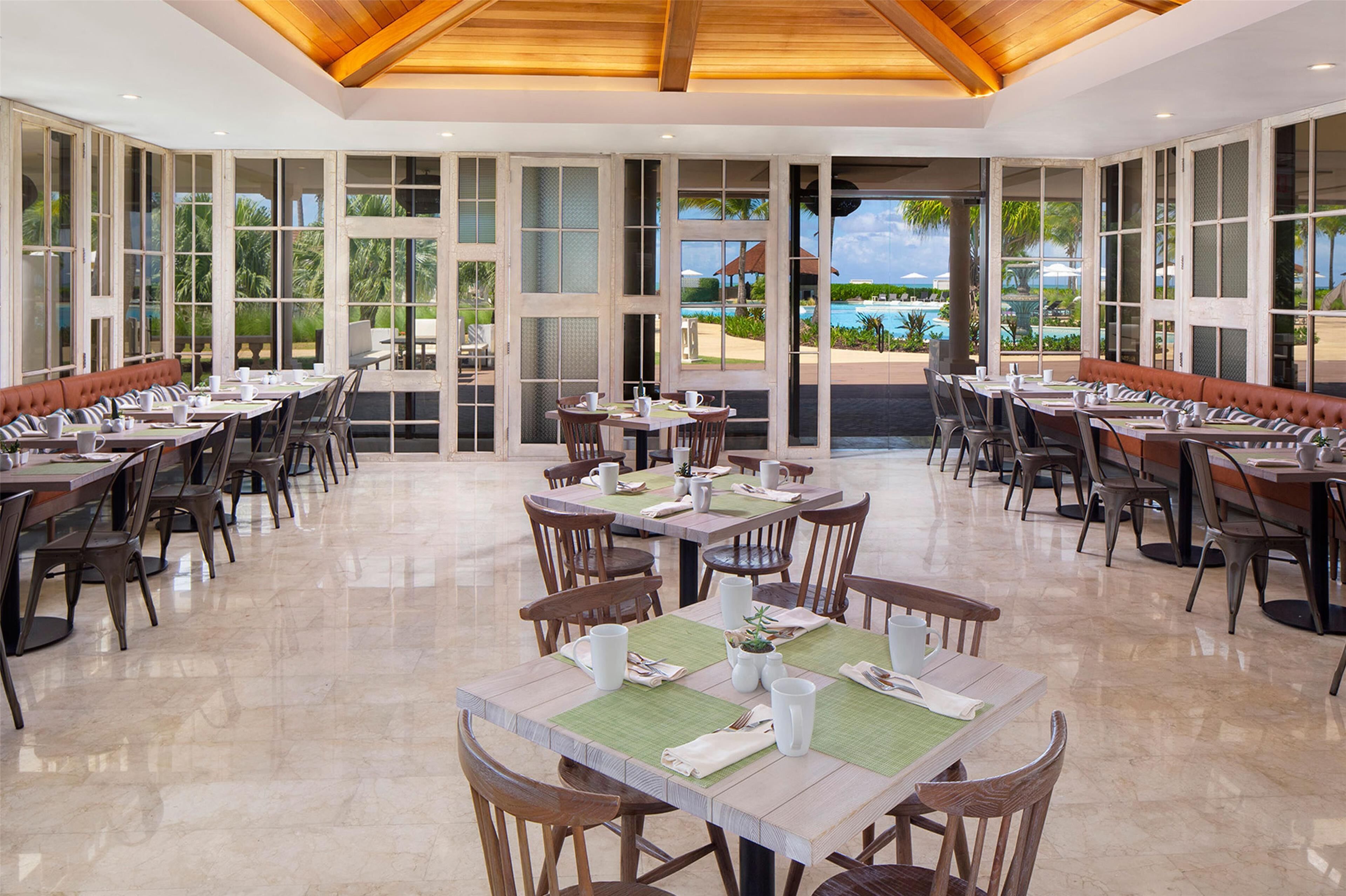 A spacious dining area with multiple tables and chairs, large windows offering views of a pool and greenery.