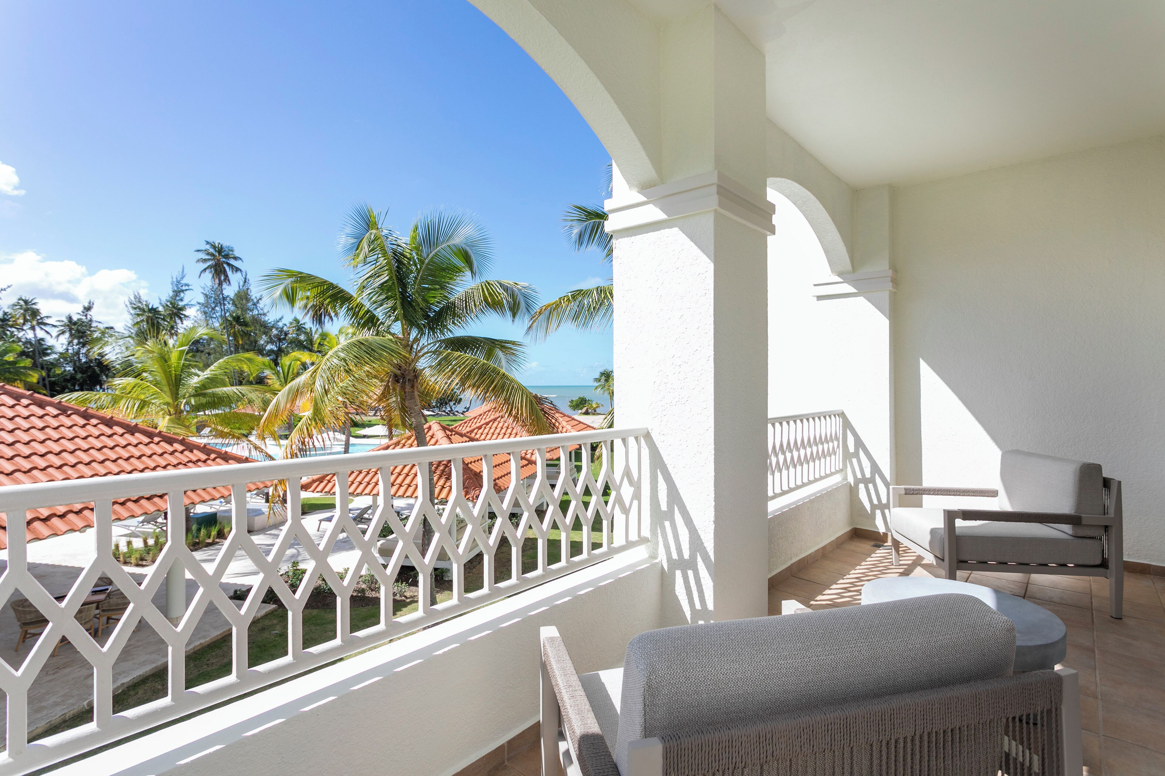 A balcony with a view of the ocean and palm trees, white railings, and a seating area with a table and chairs.