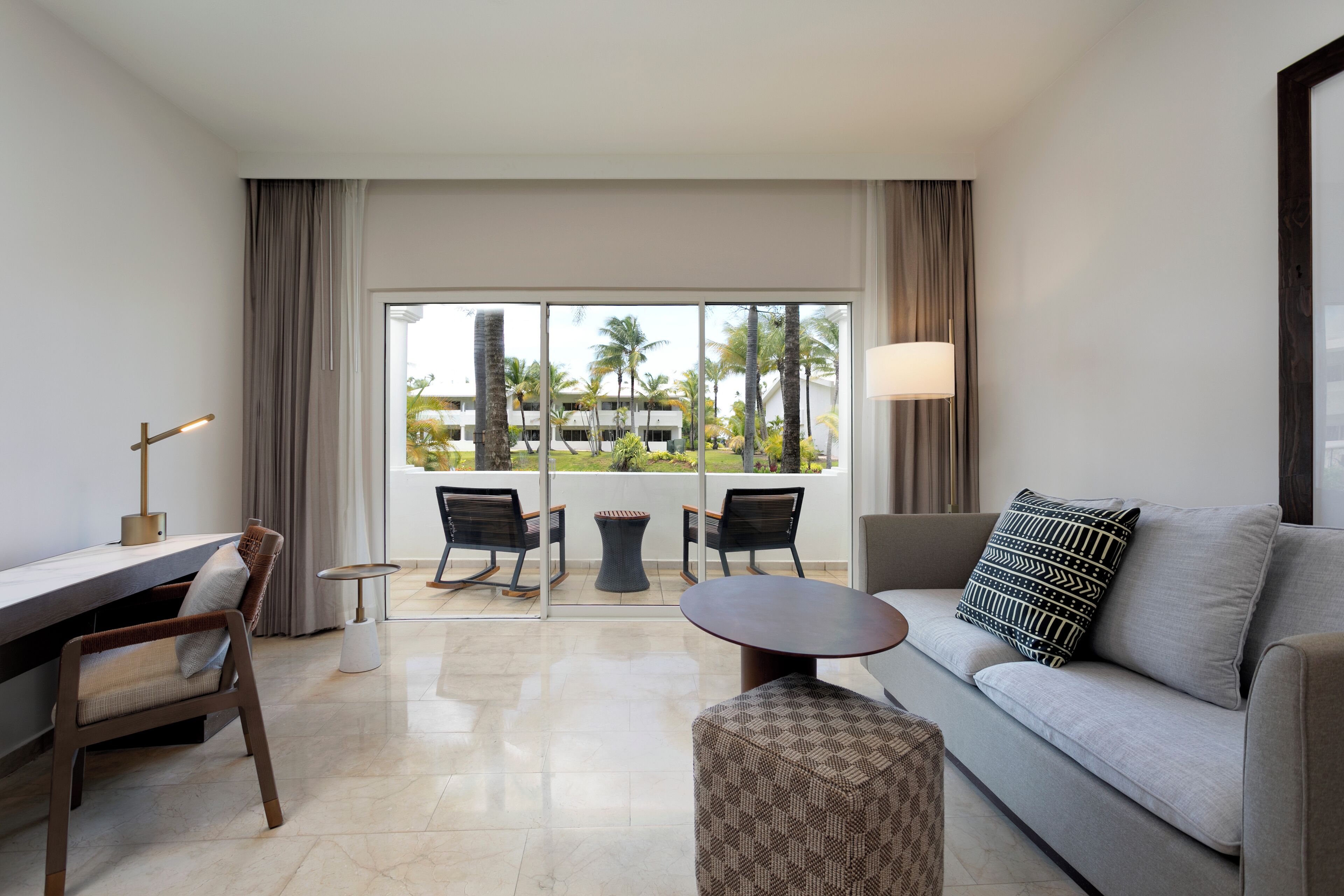 Ein modernes Hotelzimmer mit einer Couch, einem Schreibtisch, einem Sessel, einem kleinen Tisch und Blick auf Palmen durch große Fenster.