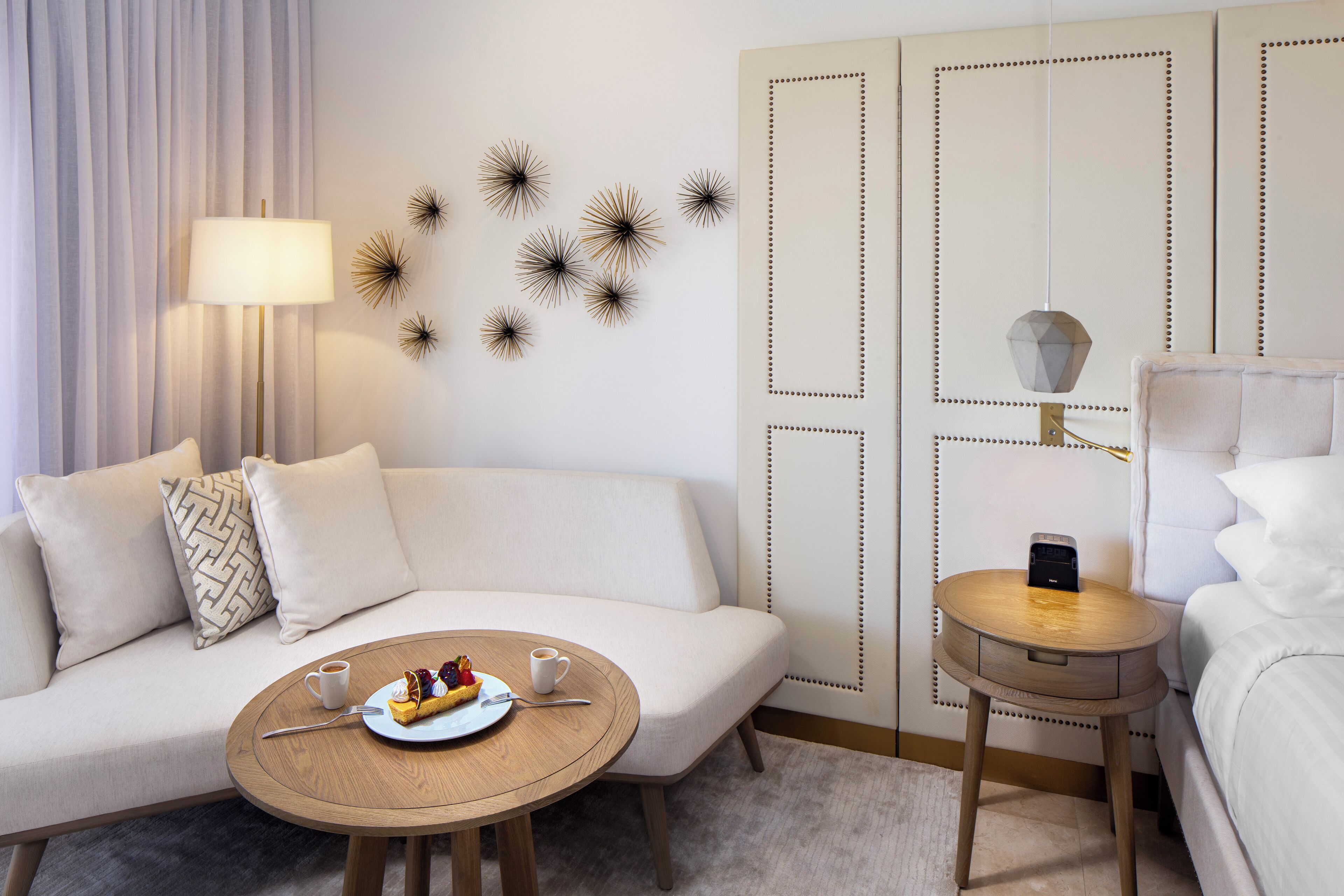 Quarto de hotel moderno com sofá, mesa redonda de madeira com prato de comida e bebidas, luminária, arte decorativa na parede e mesa de cabeceira com luminária.