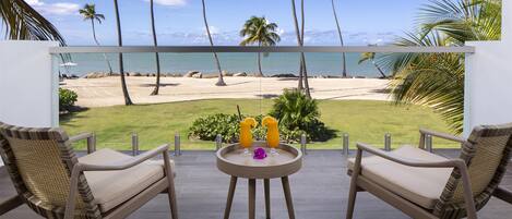 A balcony with two wicker chairs and a small round table, overlooking a beach with palm trees and a clear sky.