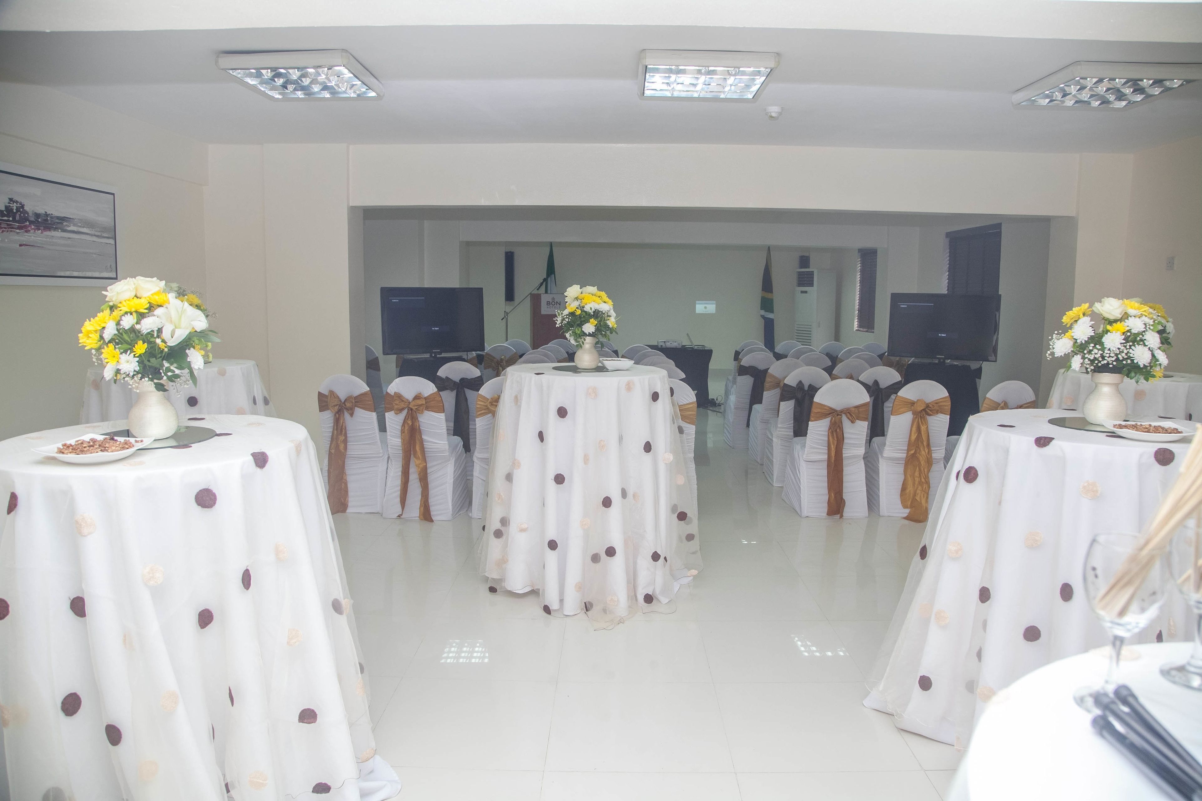 A conference room with round tables, each with a white tablecloth and a floral centerpiece. The room has a tiled floor and is equipped with ceiling lights.