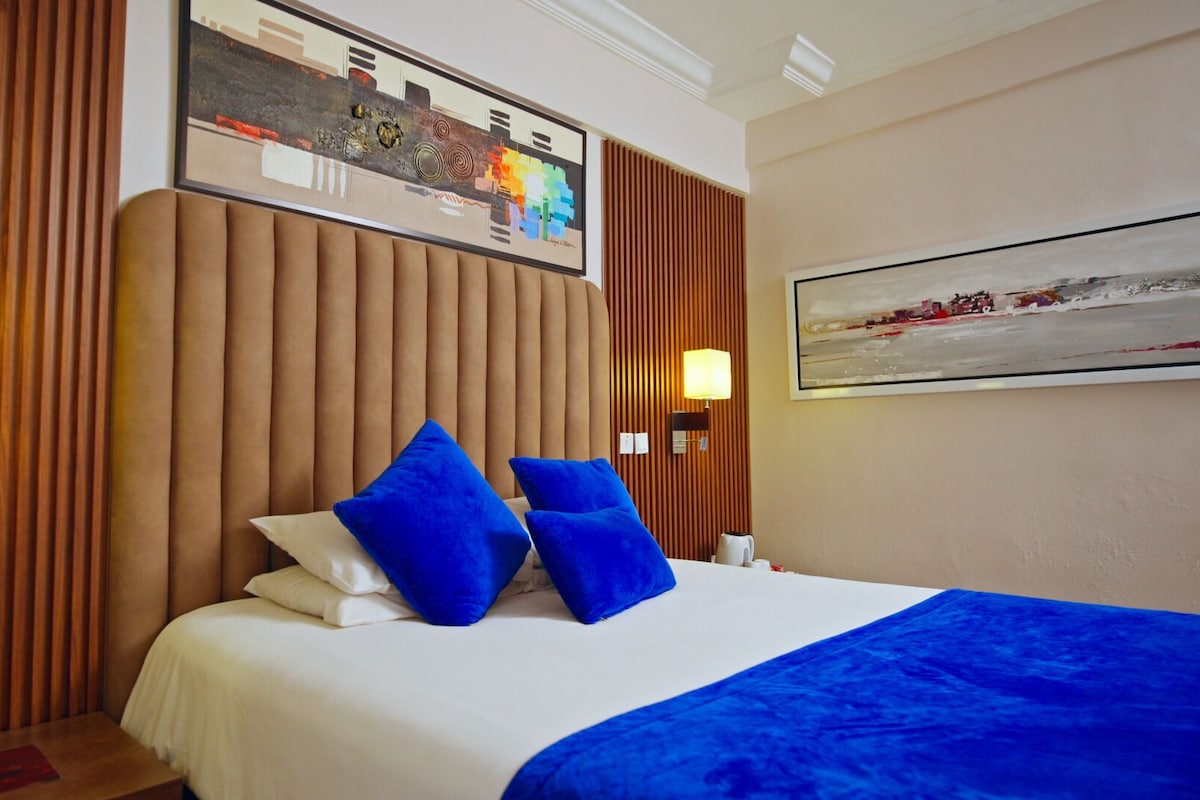 A hotel room with a large bed, two blue pillows, a white comforter, a blue throw blanket, a painting on the wall, and two wall-mounted lamps.