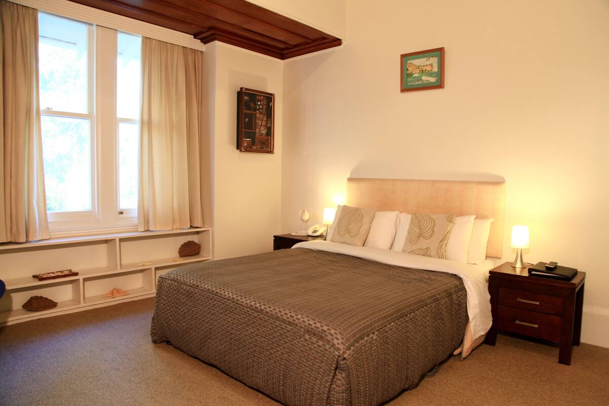 A neatly arranged bedroom with a large bed, a bedside table with a lamp, a nightstand, a window with curtains, and a framed picture on the wall.