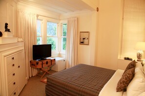 A hotel room with a bed, a TV on a wooden stand, a small desk, and a window with curtains.