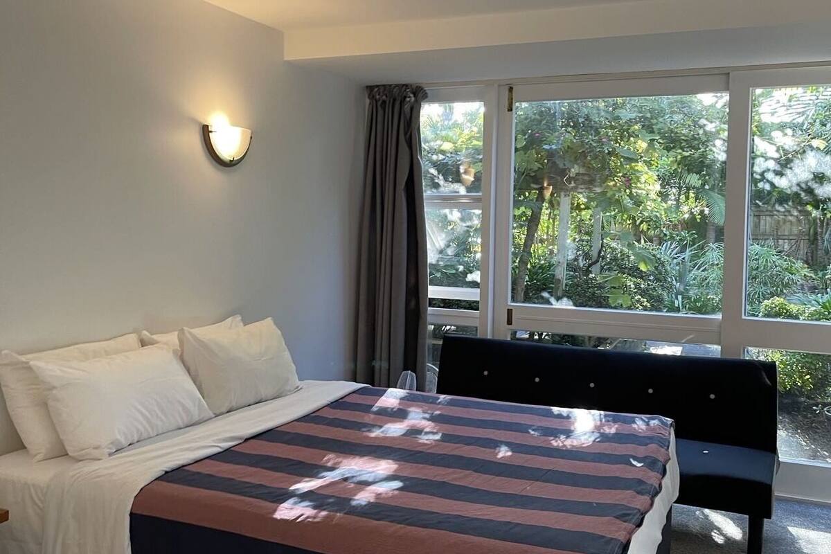 A modern bedroom with a large bed, a bedside lamp, and a view of greenery outside.