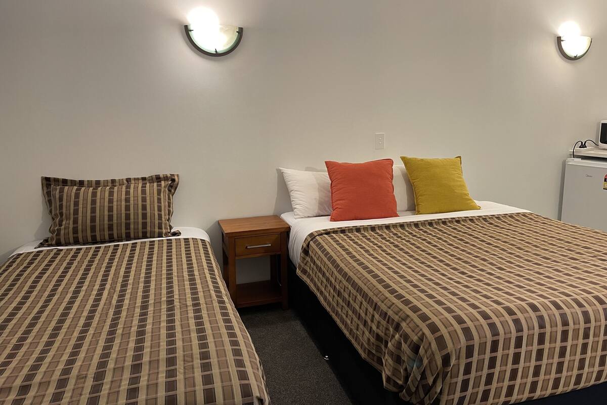 A hotel room with two beds, each with a checkered bedspread, a nightstand, a small wooden drawer, and a refrigerator.
