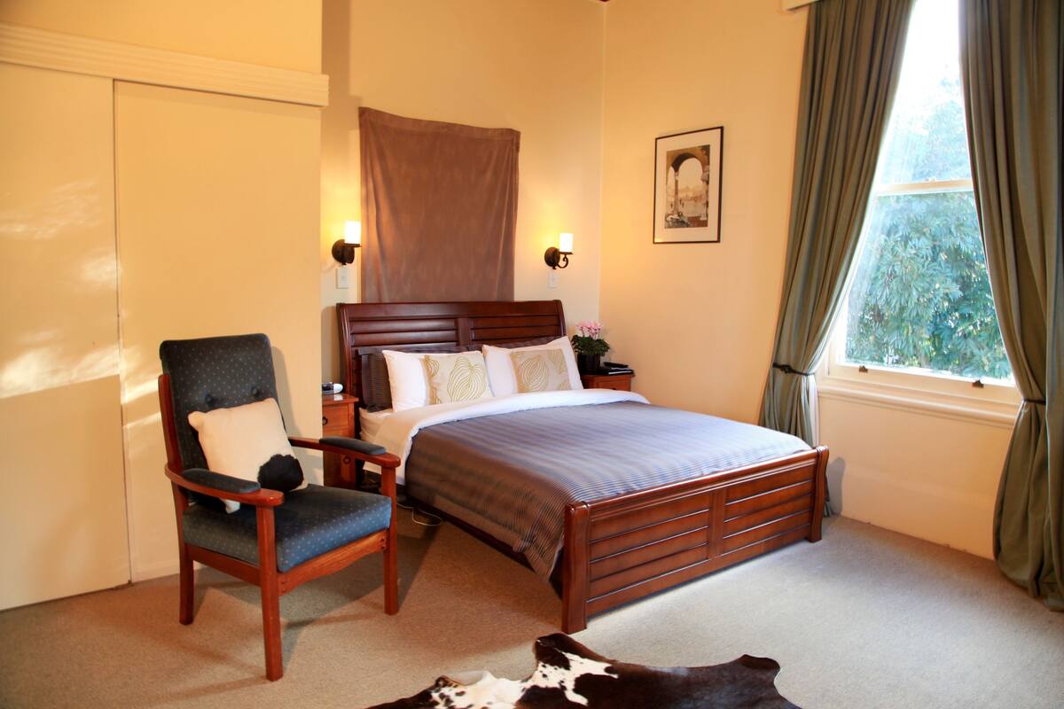 A bedroom with a wooden bed, a chair, a nightstand, and a window with curtains.