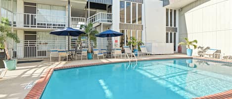 A swimming pool with lounge chairs and umbrellas, surrounded by a building with balconies and large windows.