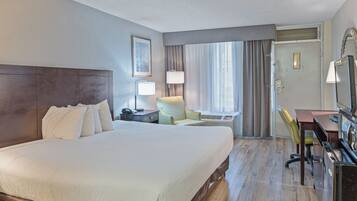 Habitación de hotel con una cama grande, un escritorio con silla, una lámpara y una ventana con cortinas.