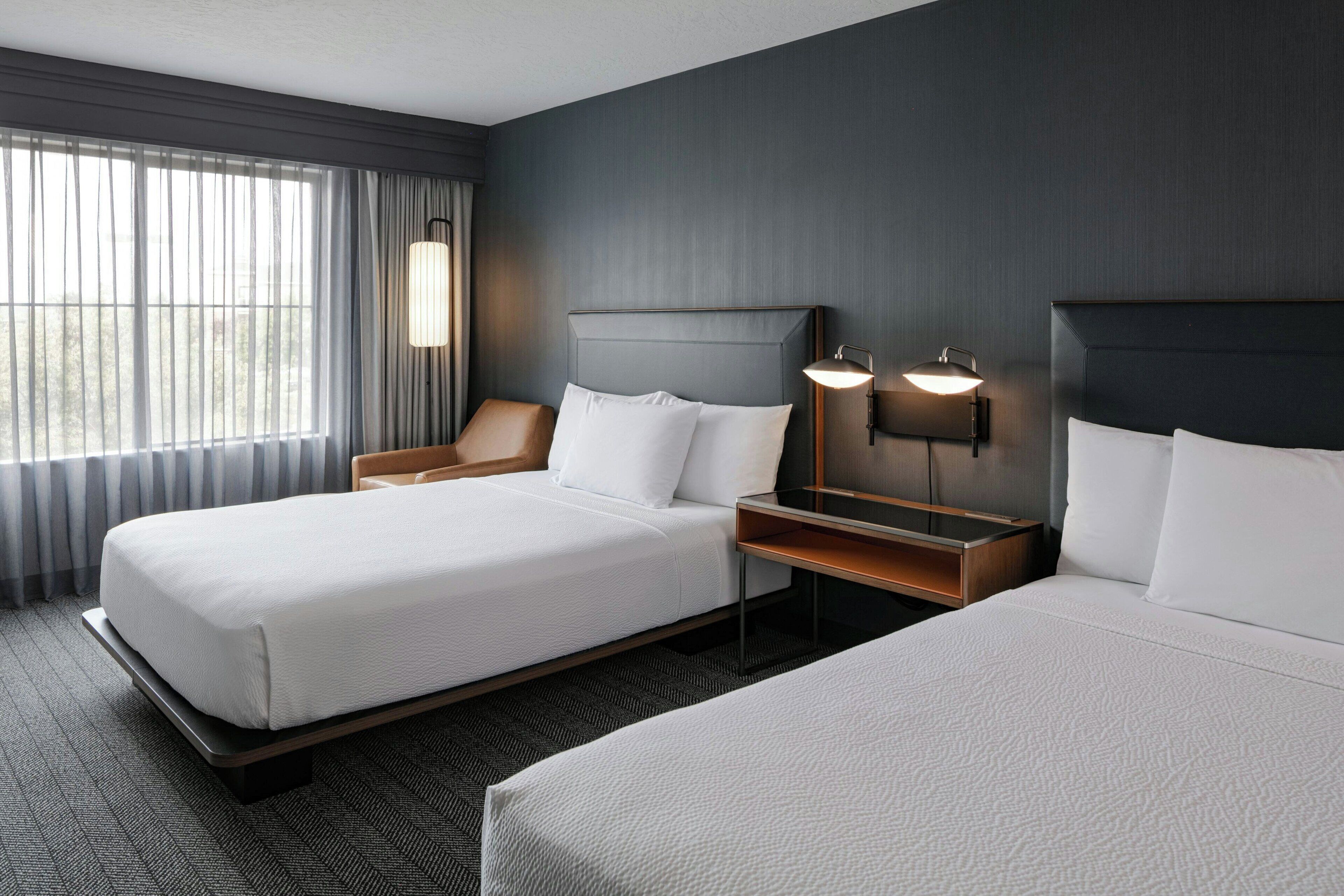A hotel room with two beds, a nightstand, a lamp, and a window with curtains.