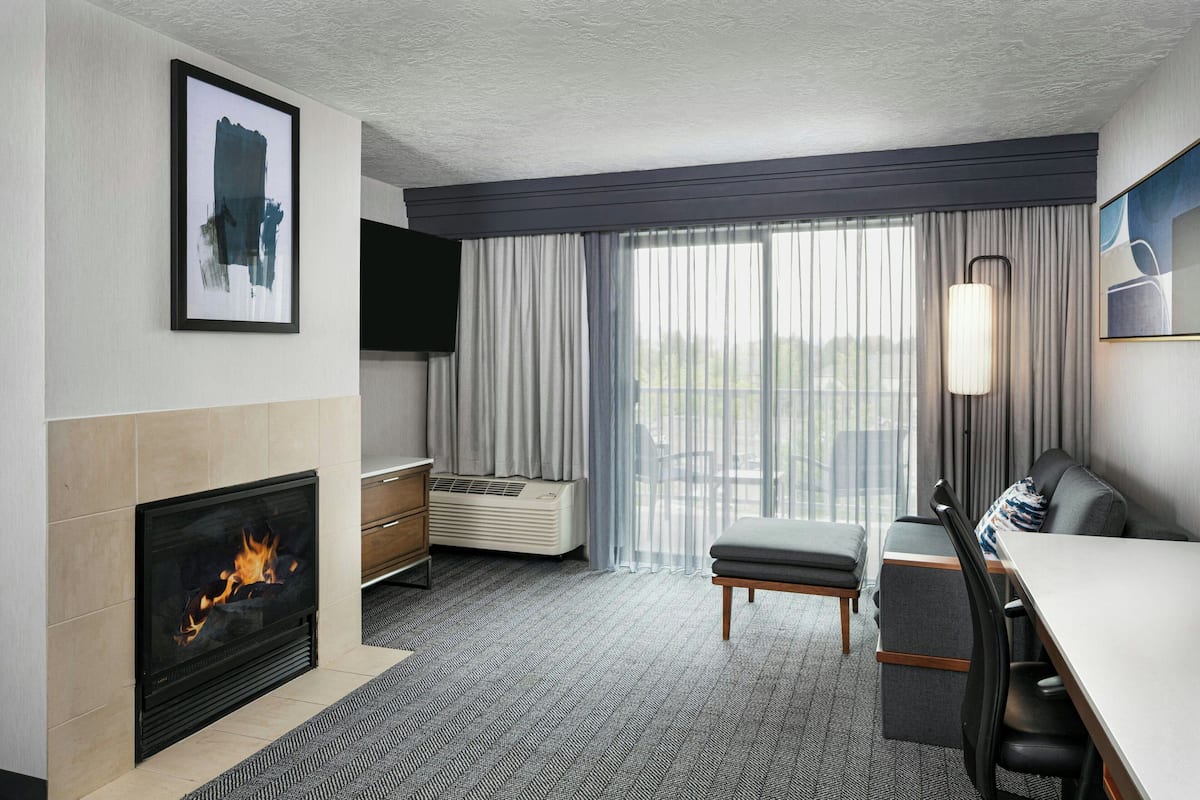 Habitación de hotel moderna con chimenea, televisor de pantalla plana, zona de comedor y un gran ventanal con cortinas.