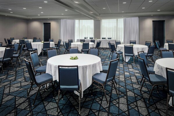 A conference room with round tables and chairs, large windows with curtains, and a carpeted floor.
