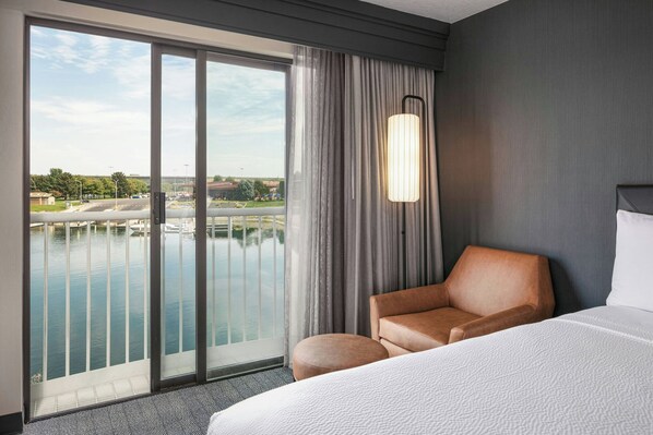 Habitación de hotel con un amplio ventanal corredera que da a una zona de piscina, una cama, un sillón y una lámpara moderna.