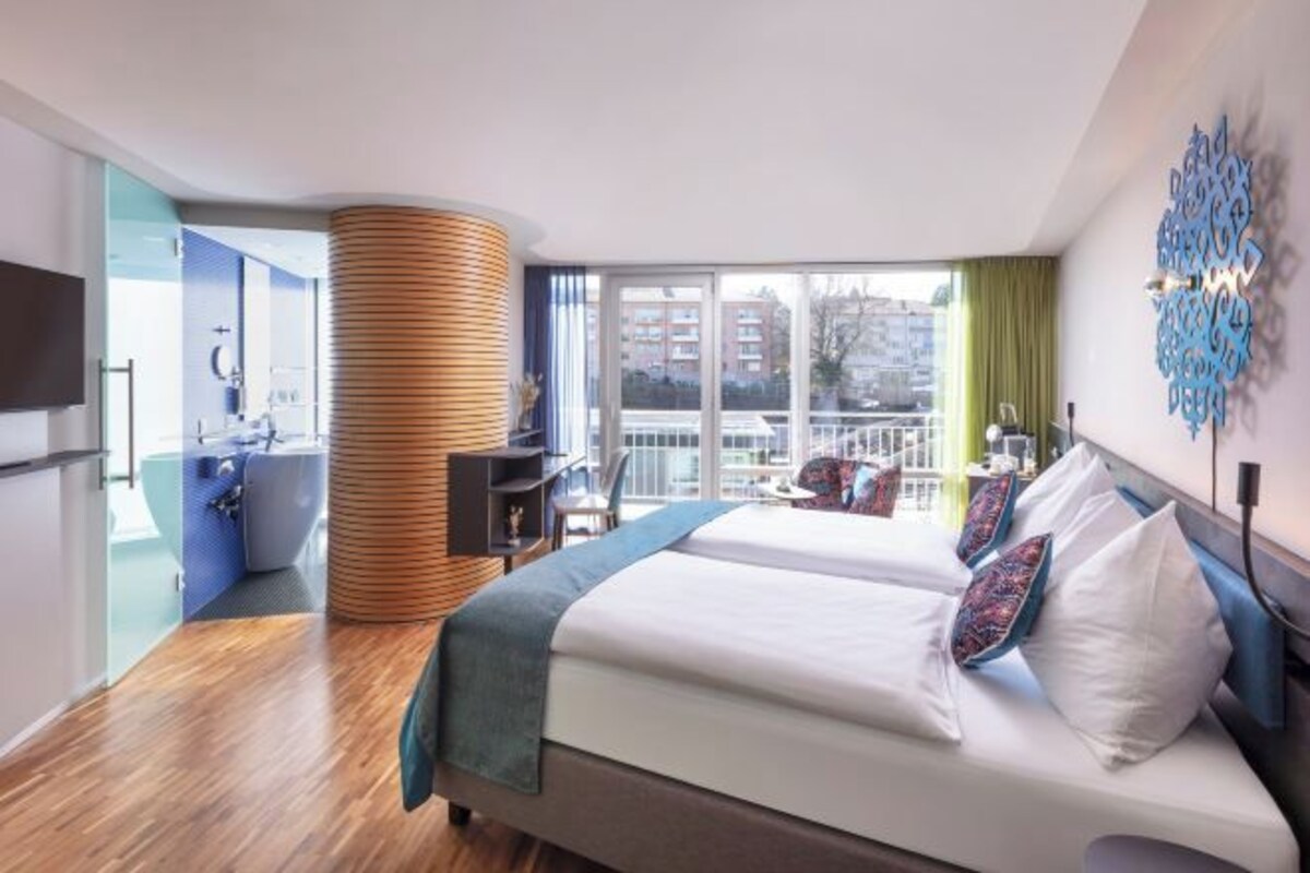 Ett modernt hotellrum med en stor säng, ett skrivbord och utsikt över staden genom stora fönster.