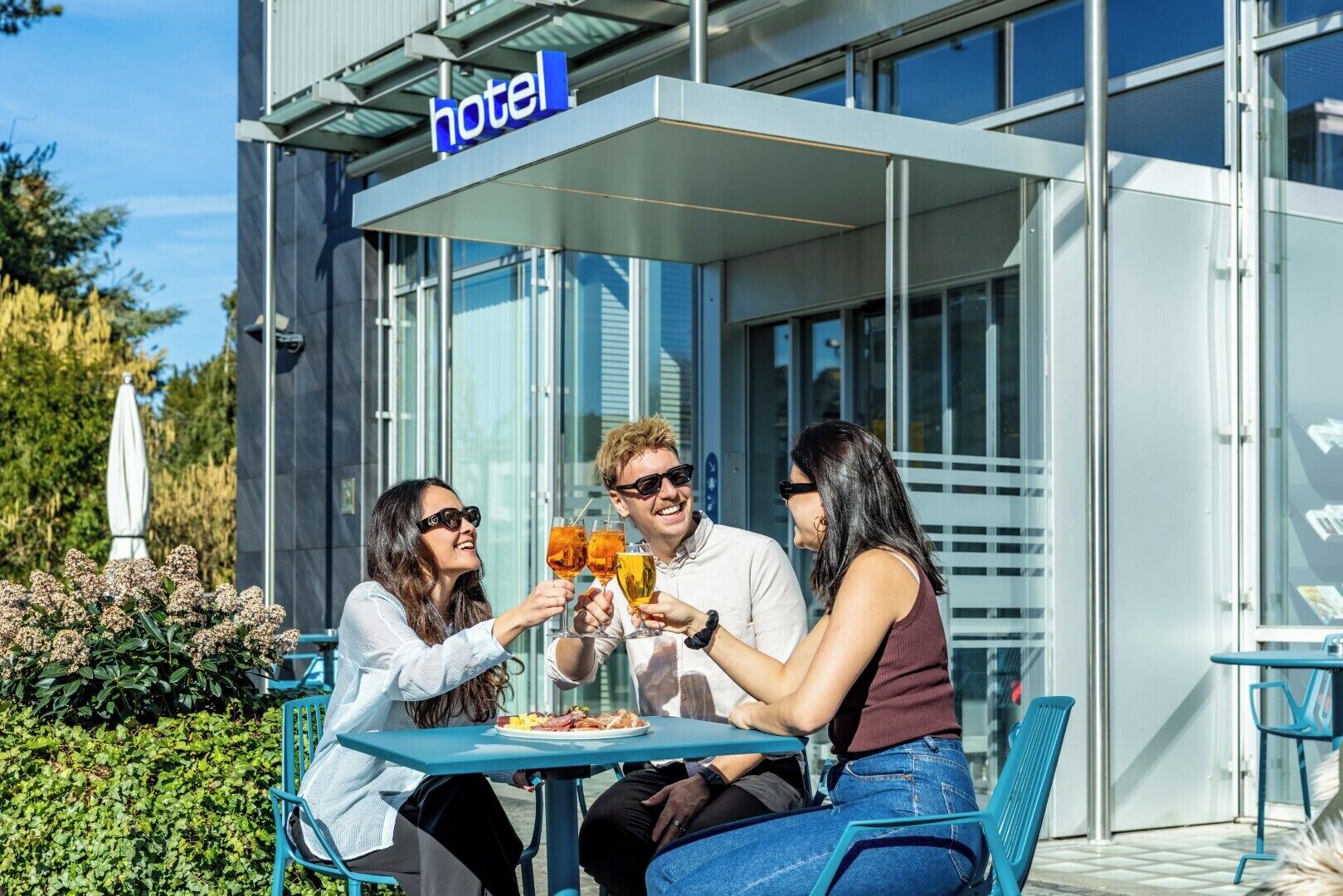 A modern hotel exterior with a glass facade, outdoor seating, and a clear sky.