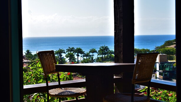 A balcony with wicker chairs and a table, overlooking a coastal view with palm trees and the ocean. - Hibiscus Beach Pousada (Búzios)