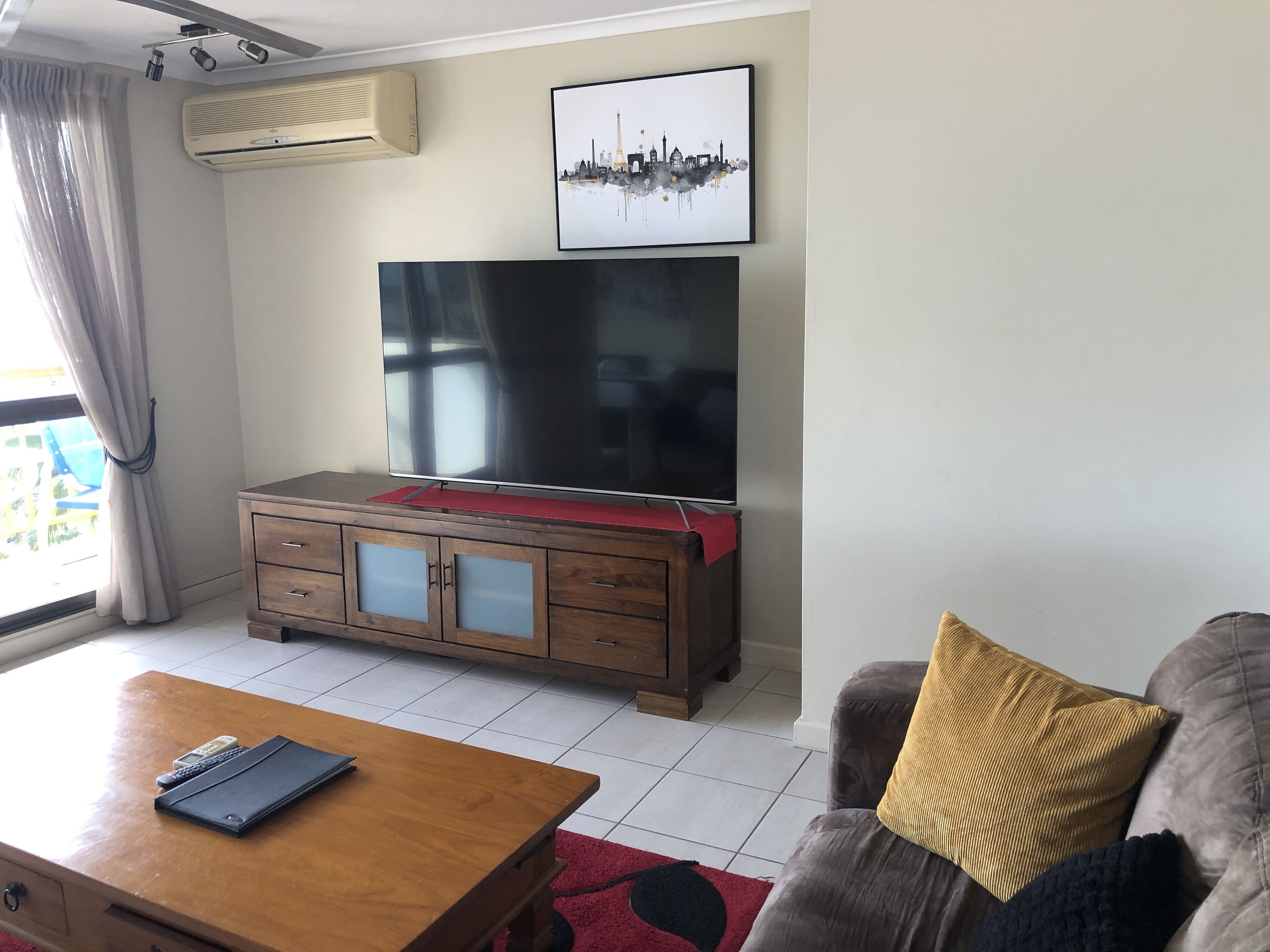 Een woonkamer met een flatscreen televisie op een houten kast, een salontafel met afstandsbedieningen en een bank met kussens.