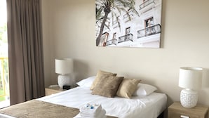 Una habitación de hotel con cama, dos mesitas de noche, ventilador de techo, un ventanal con cortinas y un cuadro colgado en la pared.