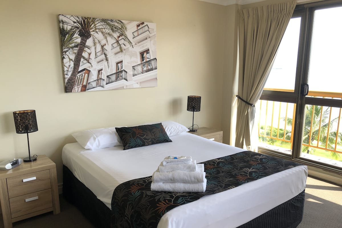 A hotel room with a large bed, two bedside tables with lamps, a painting of a building and palm trees, and a view of greenery through the window.