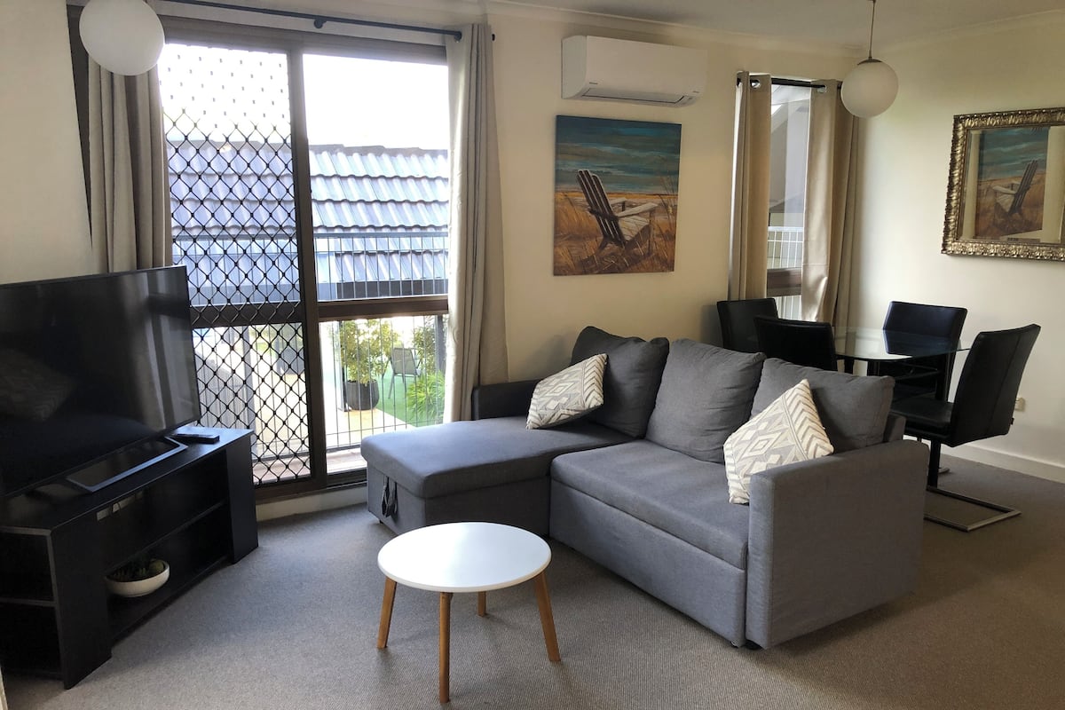 A living room with a grey sofa, a TV, a small white table, and a balcony with a view of a building with blue tiles.