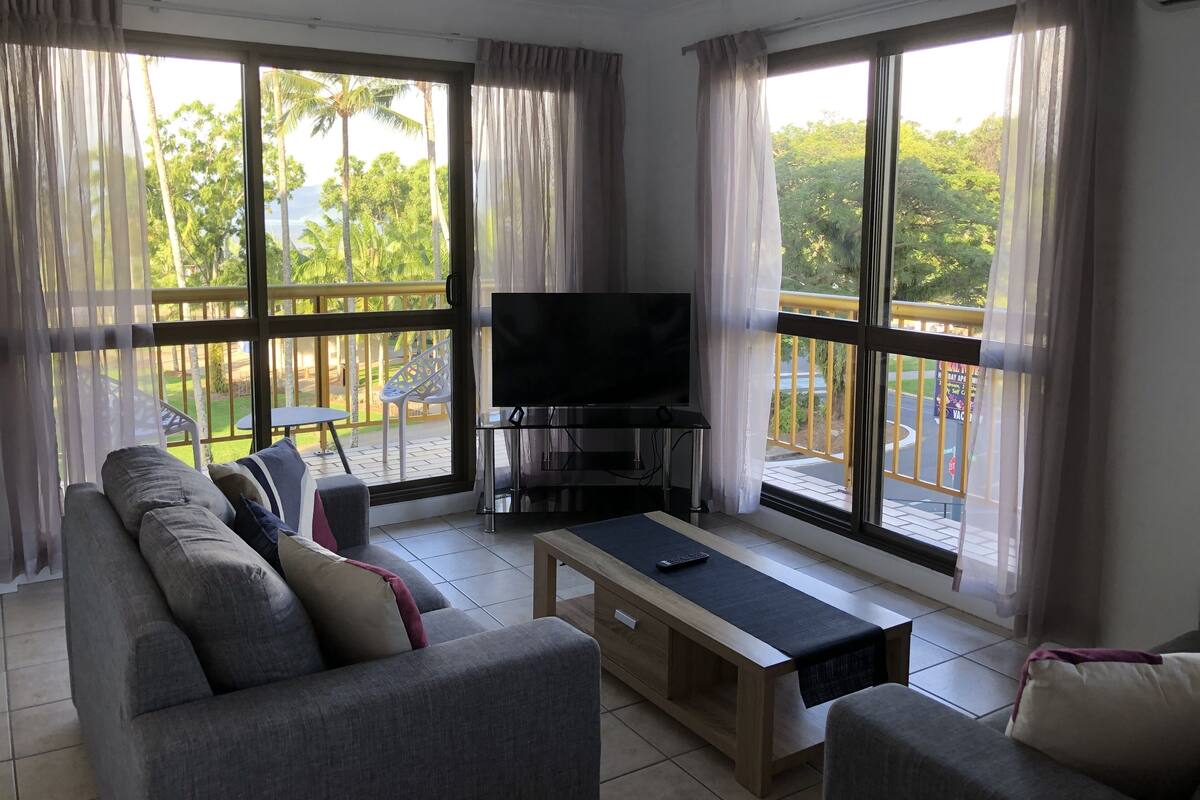 A living room with a sofa, a coffee table, a TV, and large windows with curtains.